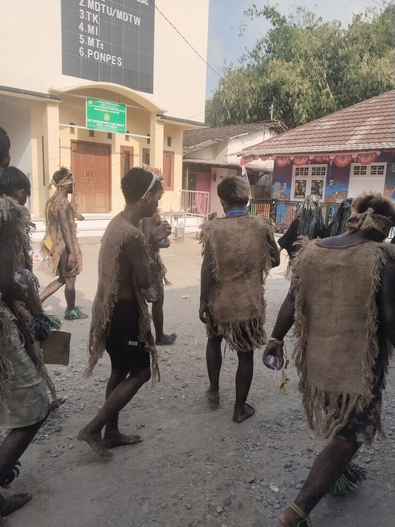 In Jembayat, Indonesia, a group of individuals are walking on a dirt path in front of a building with a sign that reads "TPQ PERGURUAN NURUL ULUM JEMBAYAT". The individuals are wearing traditional, rough-spun clothing made of what appears to be burlap or hessian, with fringed edges. Some have their faces and bodies smudged with dark material. The building behind them has a light-colored facade with wooden doors and windows. To the right of the building, there are more structures with tiled roofs, one of which is decorated with colorful murals featuring cartoon-like figures. The environment appears to be outdoors, with trees and vegetation visible in the background. The scene suggests a cultural or traditional event is taking place.