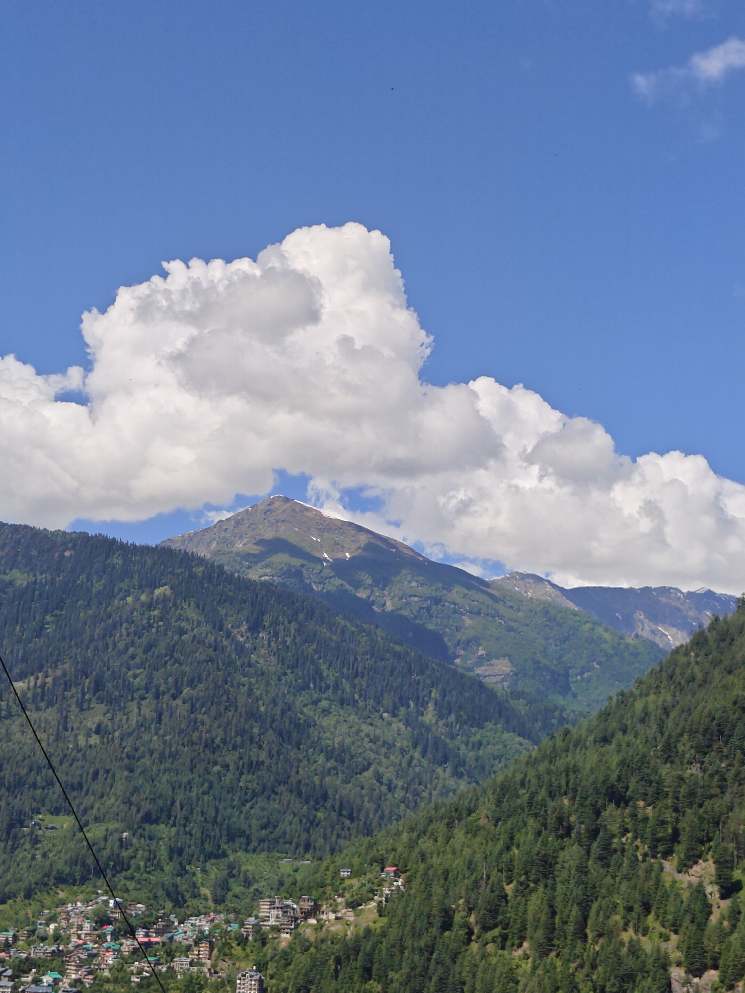 A high-angle, full outdoor shot depicts a vast mountain range covered in dense green pine trees under a bright blue sky with scattered white clouds. The mountain peaks in the distance show patches of snow. In the lower left quadrant, a village is nestled on the hillside, characterized by numerous buildings with colorful roofs. A thin black cable runs diagonally from the upper left corner down towards the village. The scene captures the natural beauty of the Vashisht, India region, characterized by its mountainous terrain and the presence of a small settlement amidst the lush greenery.