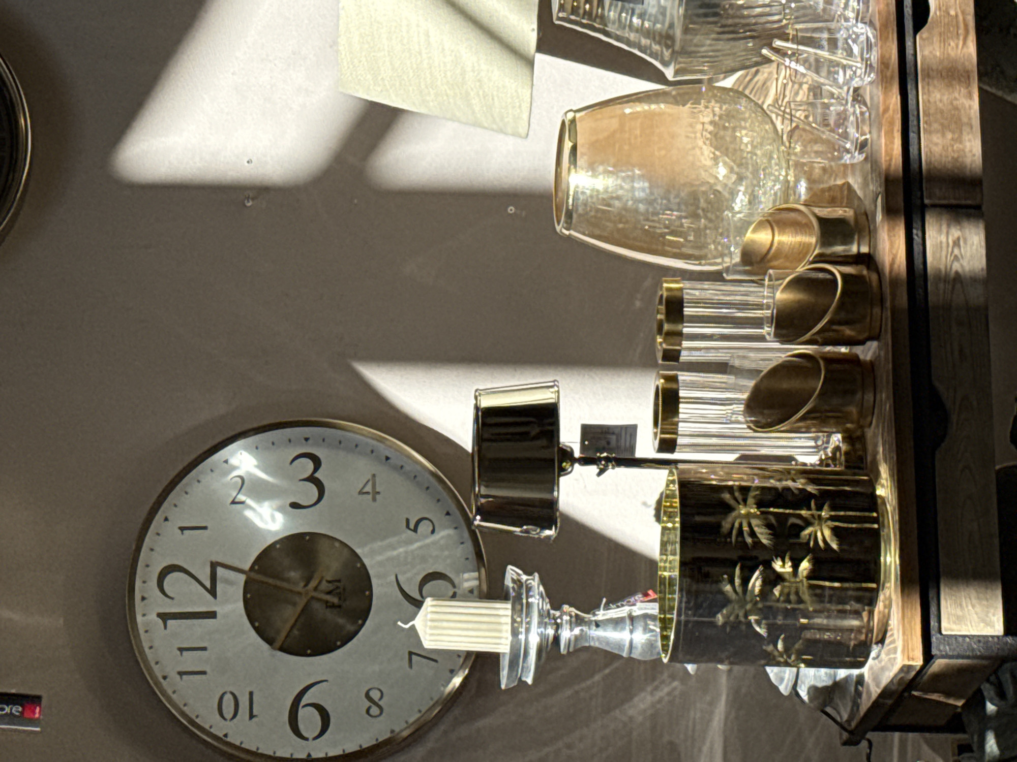 The image depicts a display of decorative objects on a wooden table, likely in a retail setting, possibly in Bad Sachsa, Germany. Dominating the background is a large, circular wall clock with Roman numerals, indicating the time as approximately 12:08. To the left of the clock is a tall, silver-toned candlestick holding a cream-colored, fluted pillar candle. Adjacent to the candle holder is a small, dark lampshade with a metallic finish.
In the foreground, a black cylindrical object adorned with a pattern of gold palm trees sits on the table. To its right are several clear glass cylinders with gold rims, some appearing to be decorative holders. Further to the right, a large, amber-toned glass vase with a textured surface is visible, and behind it, a tall, clear glass decanter or vase with vertical ridges. Several small, clear glasses, some with stirrers, are arranged in front of the amber vase.
The lighting suggests a sunlit environment, with strong shadows cast across the wall behind the objects, indicating the presence of a window or light source to the right. The overall impression is one of an interior decorative display.