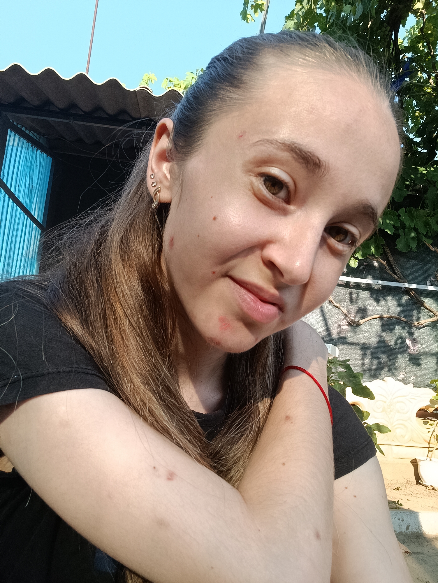 A young woman is taking a selfie outdoors, in Vypasne, Ukraine, on a sunny day. She has light brown hair pulled back and is wearing a black t-shirt. She has several piercings in her left ear and a red string bracelet on her right wrist. Her face has some redness and blemishes. The background shows a corrugated roof, some green foliage, and a textured wall.