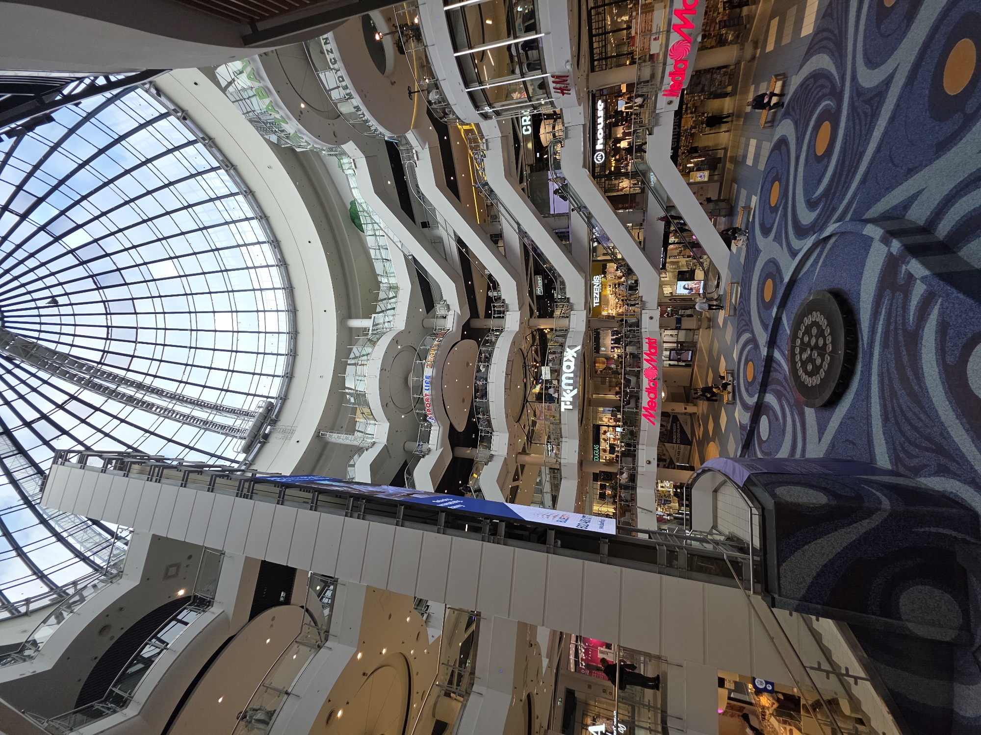 The image depicts the interior of Złote Tarasy, a shopping mall located in Warsaw, Poland. The view is oriented upwards, showcasing the mall's multi-level structure and its distinctive glass dome roof. Several escalators connect the different levels. Storefronts of various brands are visible, including "H&M," "CROPP," "HOUSE," "TK MAXX," and "Media Markt." The floor is decorated with a blue and orange patterned design. People can be seen walking and shopping on different levels.