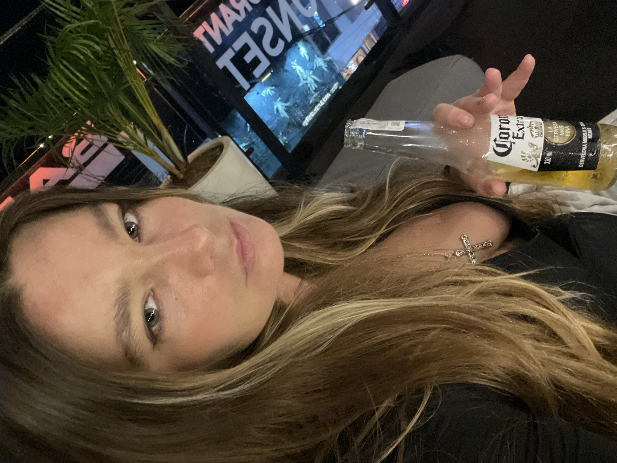 The image is a selfie taken by a woman with long, blonde-streaked brown hair. She is wearing a black top and a silver cross necklace. Her head is tilted, and she is looking at the camera with a neutral expression. In her right hand, she is holding a nearly empty bottle of Corona Extra beer. The bottle is sweating, indicating it's cold.
In the background, a palm plant in a white pot is visible to the left. Behind the plant and the woman, there is a window or glass surface with reflections and text. The visible text, partly obscured and mirrored, appears to read "TIAN" and "THET". There are also faint images of what look like statues or figures on the other side of the glass. The overall lighting suggests it is nighttime or indoors with artificial lighting. The context of "North Kuta, Indonesia" suggests this scene may be taking place in a bar, restaurant, or outdoor seating area, possibly with a view.