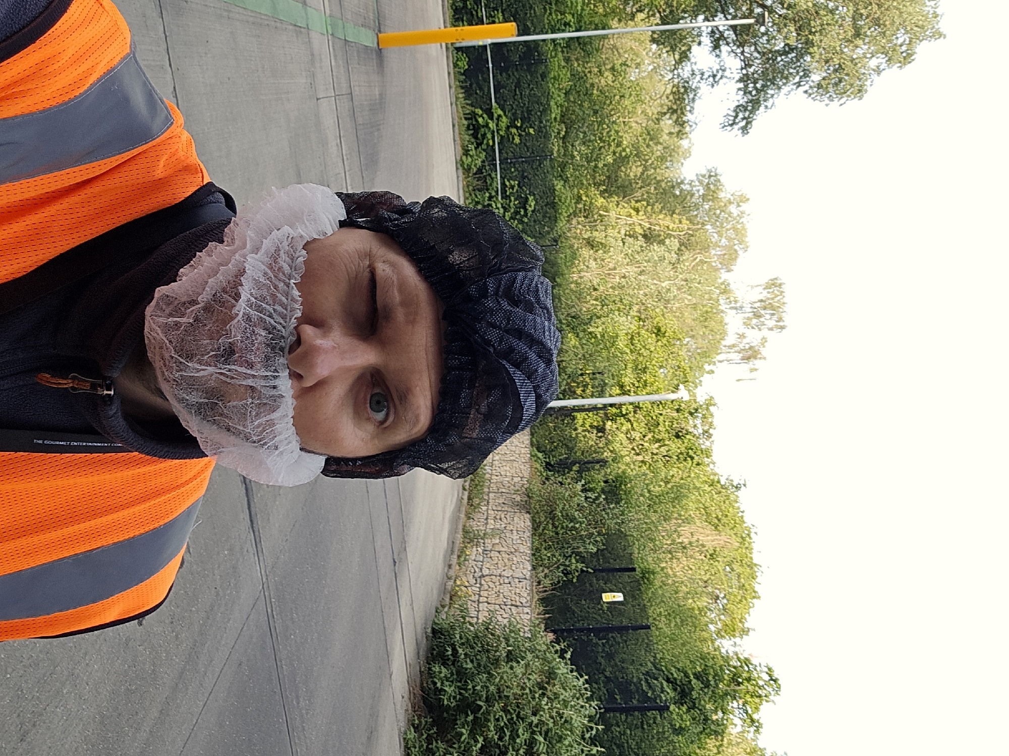 A person wearing a safety vest, hairnet, and beard net is positioned in front of a green space and sidewalk. The person is winking. The location might be a commercial area or an industrial zone, given the presence of safety gear and the surrounding landscape. While London, United Kingdom is mentioned, there are not enough details in the image to confirm that information.