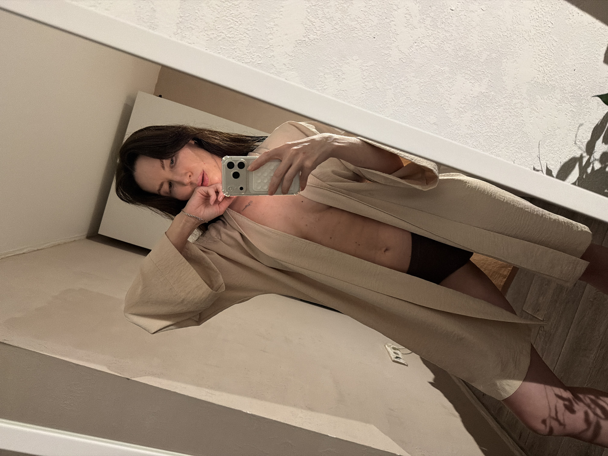 A fair-skinned woman with long, dark hair is reflected in a mirror. She is holding a smartphone in her right hand, capturing a selfie. She is wearing a light-colored, open-front garment that reveals part of her torso and the top of dark shorts. The background is a light-colored wall and a partial view of a doorway or cabinet. The photograph was likely taken indoors. The image location, Perm, Russia, is not discernible from the visual elements.
