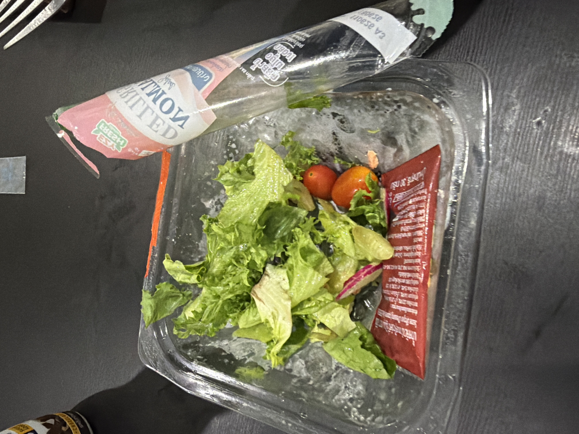 The image depicts a close-up, top-down view of a partially eaten salad in a clear plastic container on a dark, textured surface. The salad primarily consists of green lettuce, two cherry tomatoes, and thinly sliced radishes. A small, red packet, likely containing dressing, is visible beneath the lettuce. To the right of the salad container, a clear plastic wrapper with pink and white branding is present. The branding appears to be for "GRILLED SALMON." Some text in a non-English language is also visible on the wrapper. A fork with silver tines is partially visible in the upper right corner. The scene suggests a meal that has been consumed. No specific location within Baan Mae Nam, Thailand is identifiable from the visual information provided. The overall impression is of a casual, possibly take-out, meal.