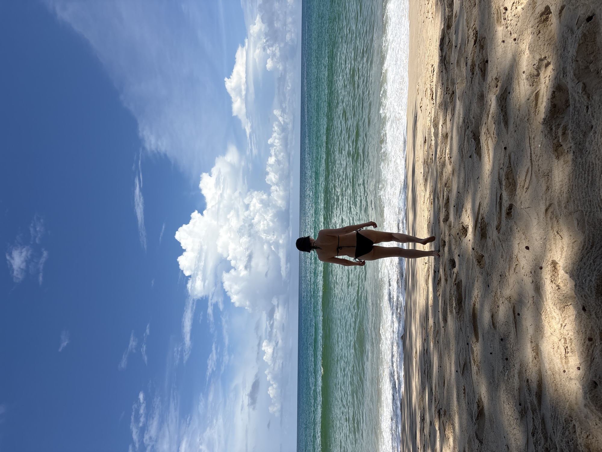 A woman wearing a black bikini and a dark cap walks towards the turquoise ocean on a sandy beach. The sky is a vibrant blue with scattered white clouds. The waves are gently breaking on the shore, creating a white foam. The sand is light brown and shows footprints. The image appears to be taken from behind the woman, looking towards the sea. The location is identified as a beach in Choeng Thale, Thailand.