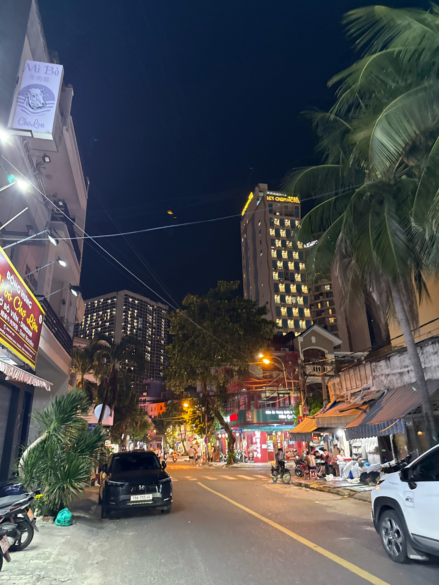 The image depicts a nighttime street scene in Nha Trang, Vietnam, likely along a commercial area given the presence of shops and hotels. On the left, a building has a sign reading "Mì Bò" with a stylized cow logo and "Cho Lon" below it. Further down the street on the left, a red and yellow sign reads "ĐÔNG HƯNG BÒ CHỢ LỚN - DIMSUM" and includes text in Vietnamese and English. A black SUV with license plate "79A-755.46" is parked on the right side of the road. Across the street, a tall building, identified by a glowing yellow sign as "LET'S CHAM HOTEL," is visible, with many illuminated windows. Palm trees with fronds are prominent, particularly on the right side of the image. Several motorcycles are parked along the sidewalk. People are seen sitting at tables outside a shop on the right side of the street, suggesting outdoor dining or a gathering. The street is paved and marked with yellow lines. The overall atmosphere is that of a lively urban environment at night, with artificial lighting from buildings, signs, and streetlights illuminating the scene.