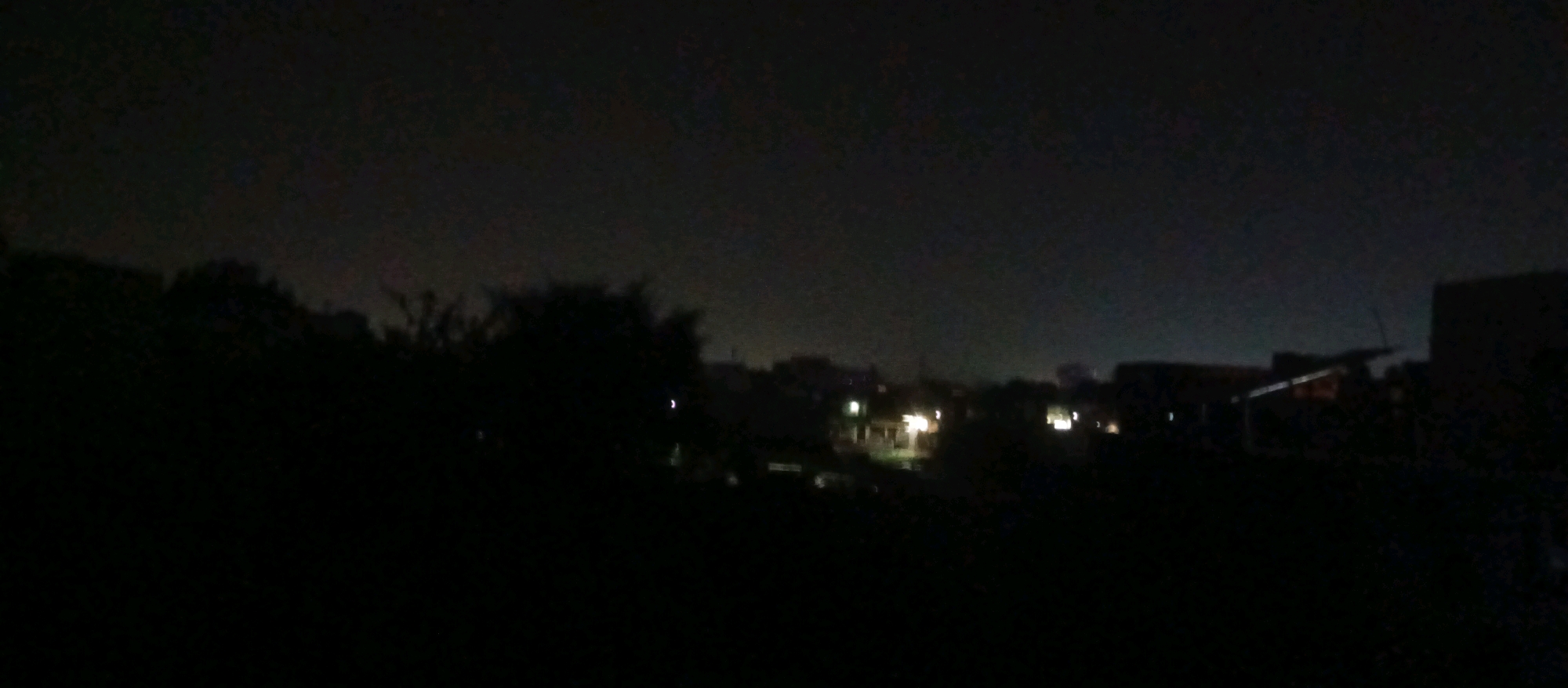 The image shows a dark, blurry landscape at night, likely a residential area in the city of Faizabad, India. Silhouettes of trees and buildings are visible against a faintly illuminated sky, possibly due to light pollution or a distant light source. Several scattered lights, appearing as small, bright dots, suggest illuminated windows or streetlights within the town. The overall impression is one of quietude and low visibility, typical of a nighttime urban or semi-urban setting. No people or specific actions are discernible due to the darkness and blurriness of the image.