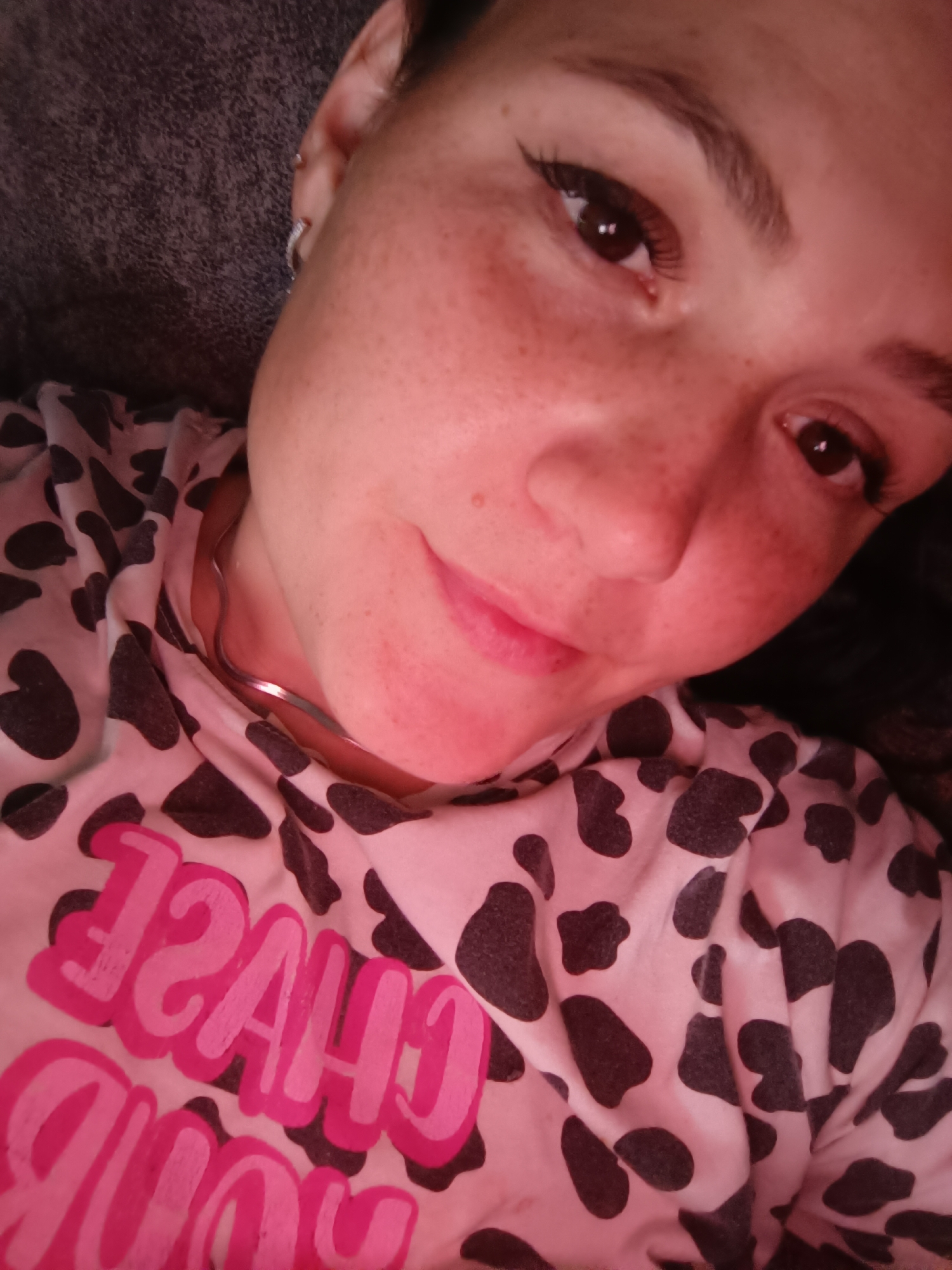 The image shows a close-up, top-down view of a young woman lying down. She has fair skin with freckles and is wearing makeup, including eyeliner and mascara. Her left ear has a small hoop earring. She is wearing a light-colored shirt with a black and white cow print pattern and bold pink text that reads "CHASE YOU". A thin, silver-toned necklace is visible at her throat. The lighting in the image is warm, casting a reddish hue. The background is dark and appears to be a soft surface, possibly a blanket or pillow. There is no indication of the city of Karsha, Russia in the image.