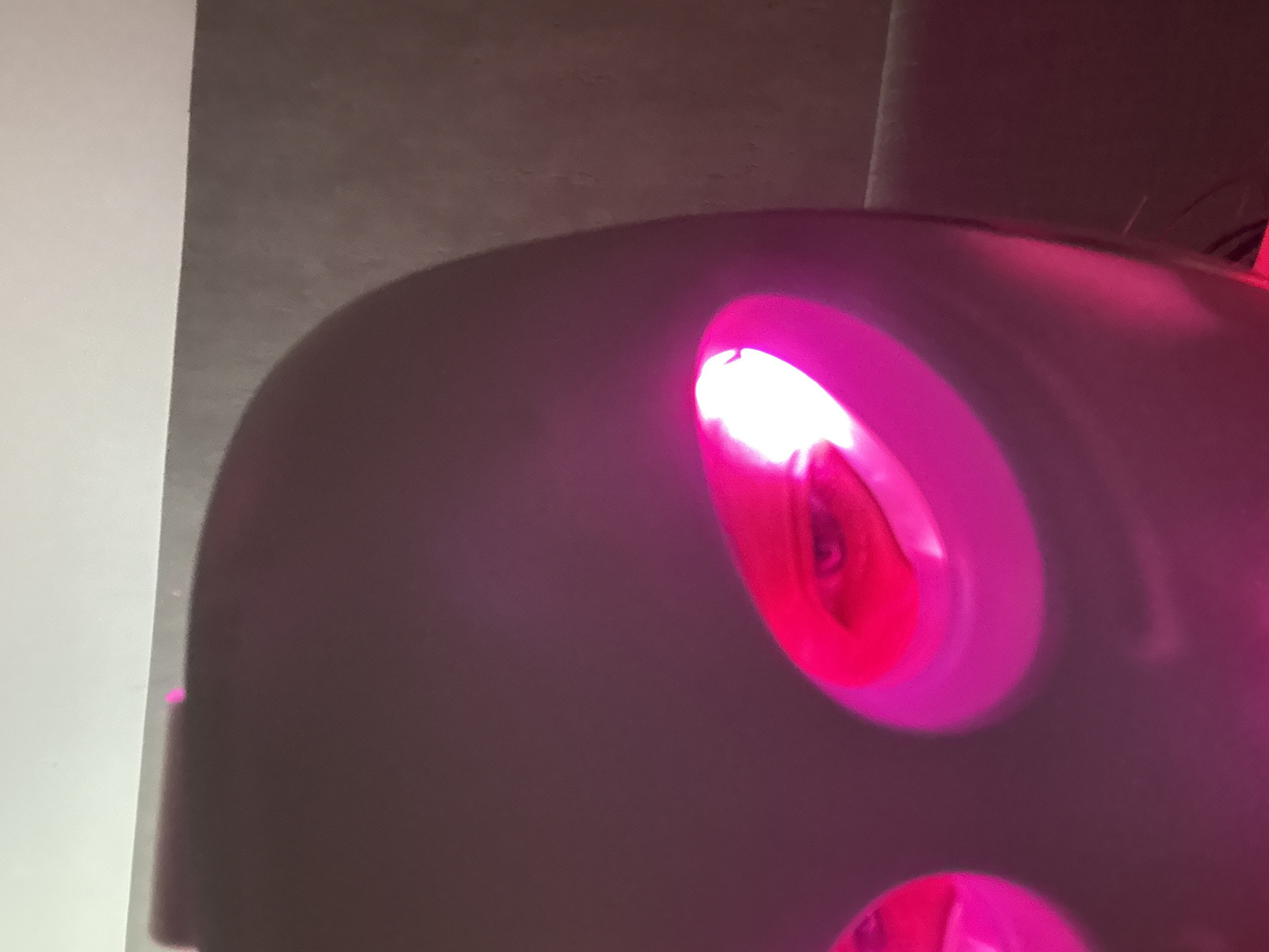 The image displays a close-up view of a dark, smooth object, likely a device, emitting vibrant magenta and pink light. The object has at least two circular light sources; one is prominently featured towards the upper right, glowing intensely bright, almost white at its center, with discernible internal textures or reflections. A second, similar light is partially visible at the bottom edge of the frame. The main body of the object is a deep, dark gray or purple hue. To the left, a sharp vertical line separates the dark object from a brightly lit, white surface, possibly a wall. The background behind the object is uniformly dark and indistinct, suggesting an indoor setting with low ambient light, likely at night, where the emitted pink light is the dominant illumination. There are no people visible, nor is there any discernible text within the image. The scene is located in San Bernardino Tlaxcalancingo, Mexico.