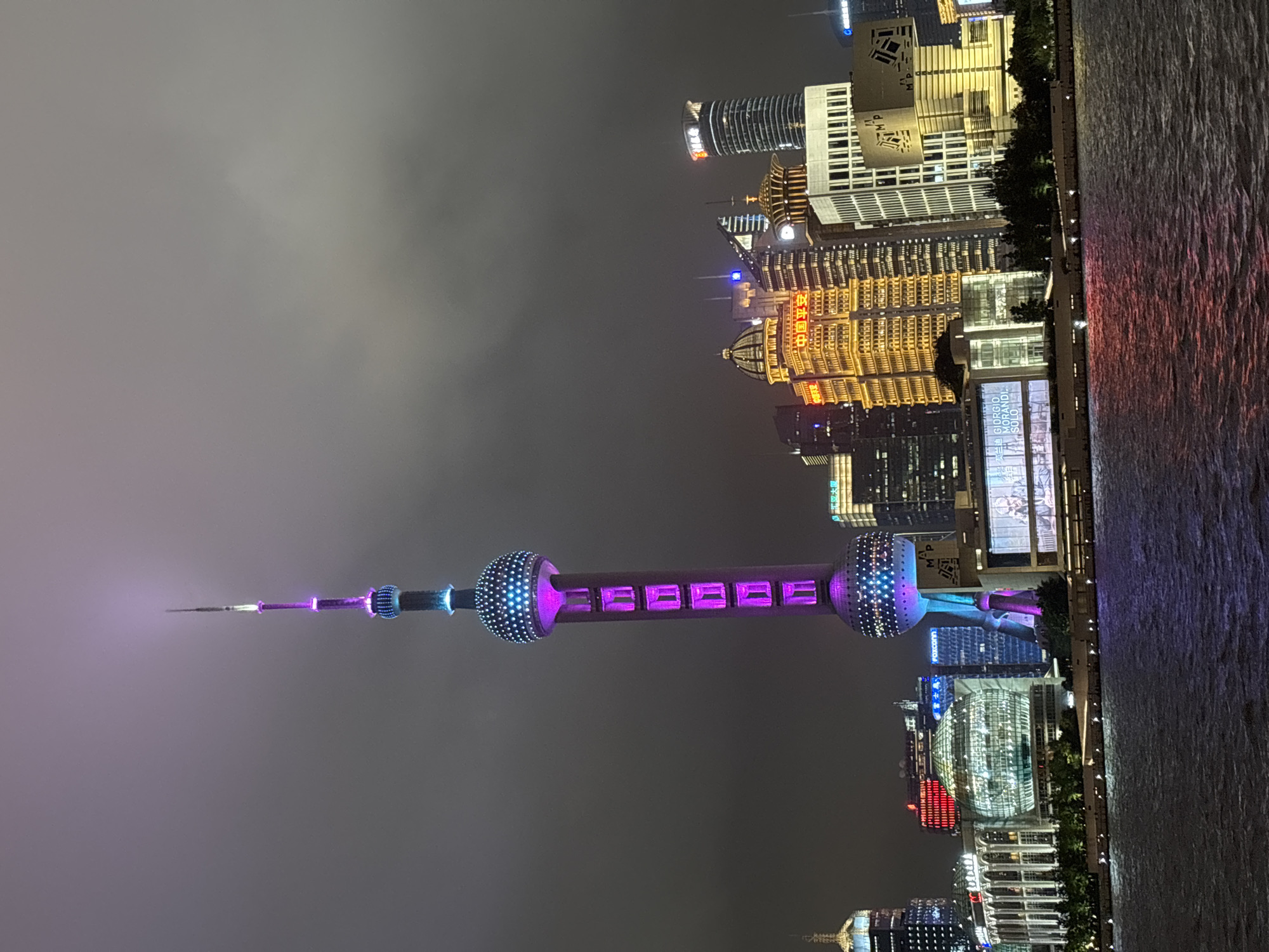 The image shows a nighttime view of the Pudong skyline in Shanghai, China, with the Oriental Pearl Tower prominently featured. The tower is illuminated with purple and blue lights, and its spherical sections are clearly visible. Surrounding the tower are various skyscrapers, many of which are also lit up with colorful lights. One building on the right has a sign that appears to be in Chinese characters. In the foreground, a body of water reflects the lights from the city. The sky is dark and overcast, suggesting nighttime. There are no people or identifiable actions depicted in the scene. The overall impression is a vibrant urban landscape at night.