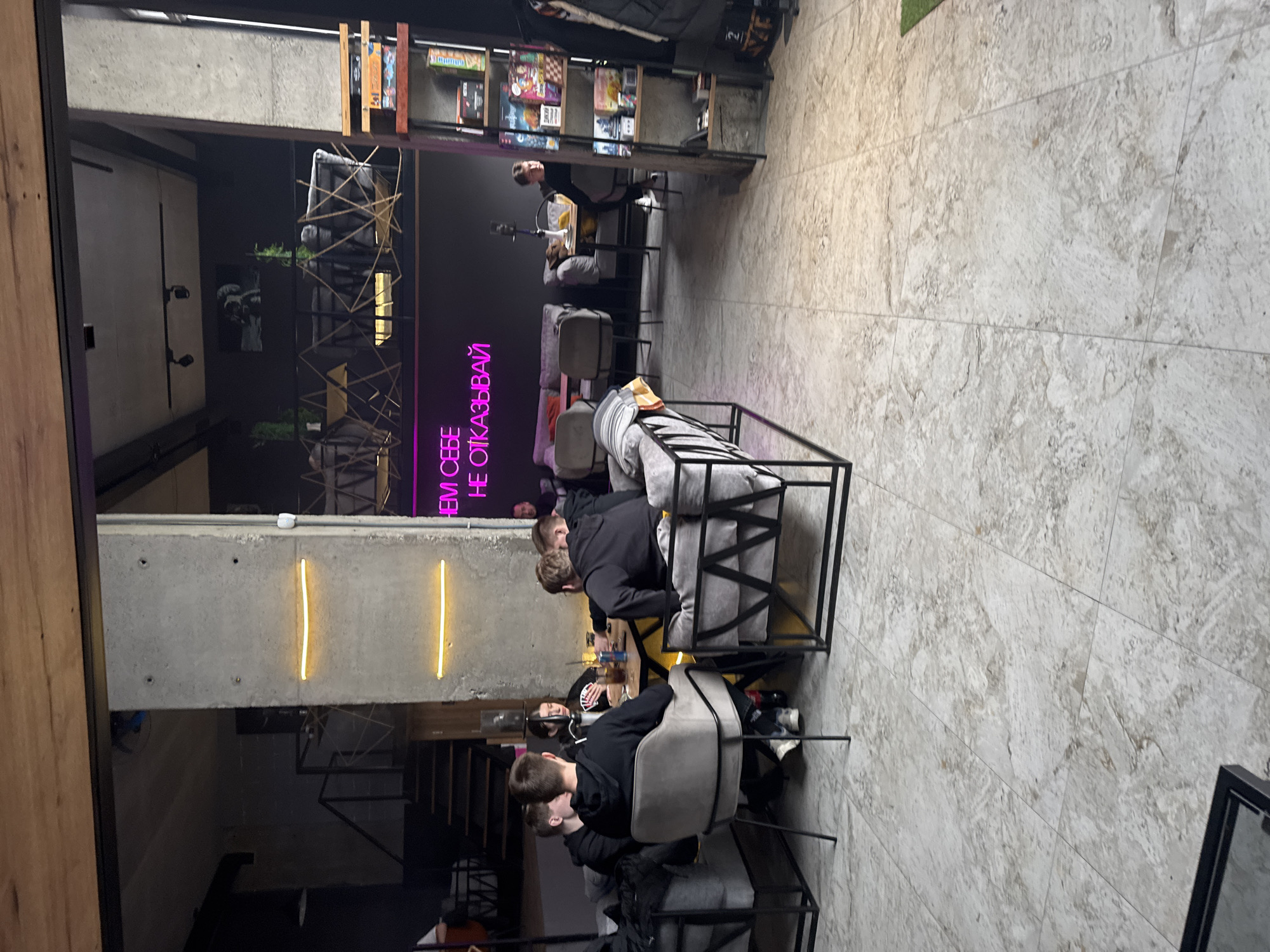 The image depicts an interior scene in an unknown location in an unknown city. Three people are seated at a table, seemingly engaged in a card game. The individuals are wearing casual attire. The table is cluttered with drinks and playing cards. The surrounding environment consists of a modern, industrial-style space. There are dark walls with a neon sign in what appears to be a different language. Additionally, there is shelving containing board games and decorative elements. The flooring consists of light-colored tiles. The overall atmosphere suggests a casual social gathering.