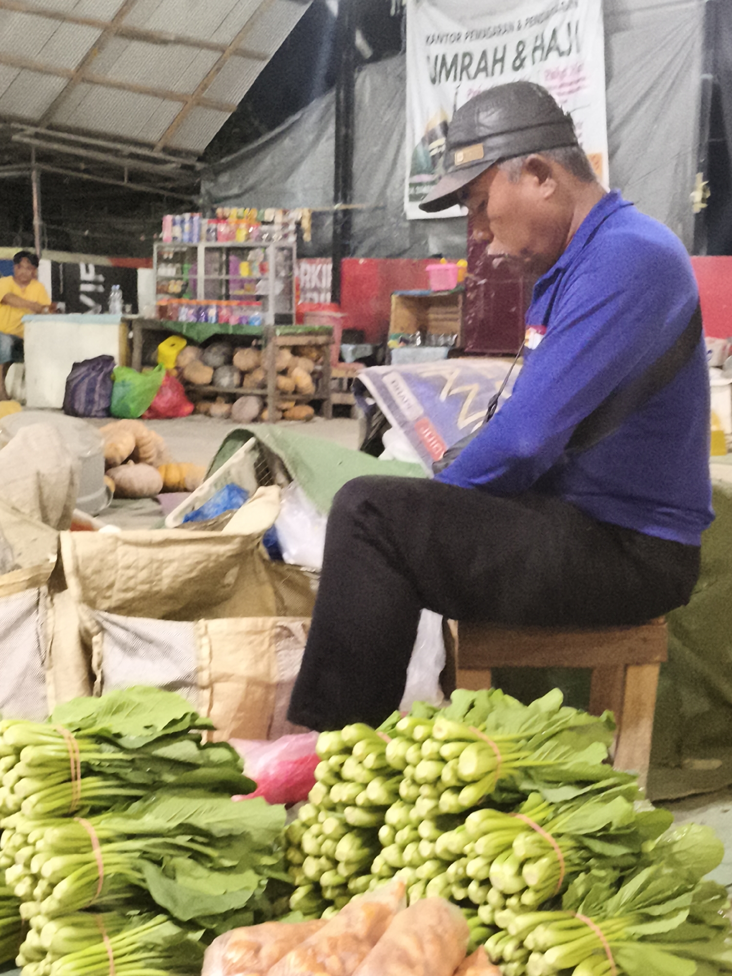 This image depicts an outdoor market scene, likely in Samarinda, Indonesia, given the context. In the foreground, bunches of green leafy vegetables, possibly mustard greens or bok choy, are stacked and bound with rubber bands. To the right, another pile of similar vegetables is visible. In the middle ground, several large, light-colored sacks are present, and some gourds or pumpkins are scattered on the ground behind them. A man wearing a blue long-sleeved shirt and dark pants, with a dark baseball cap, is seated on a small wooden stool. He is looking down at a document or book he is holding. Behind the man, a banner with Indonesian text, including "UMRAH & HAJI," is displayed, suggesting a travel agency or related service. In the background, there is a stall with shelves filled with various items, and another person in a yellow shirt is partially visible. The overall environment appears to be a bustling marketplace with goods for sale and people engaged in various activities.