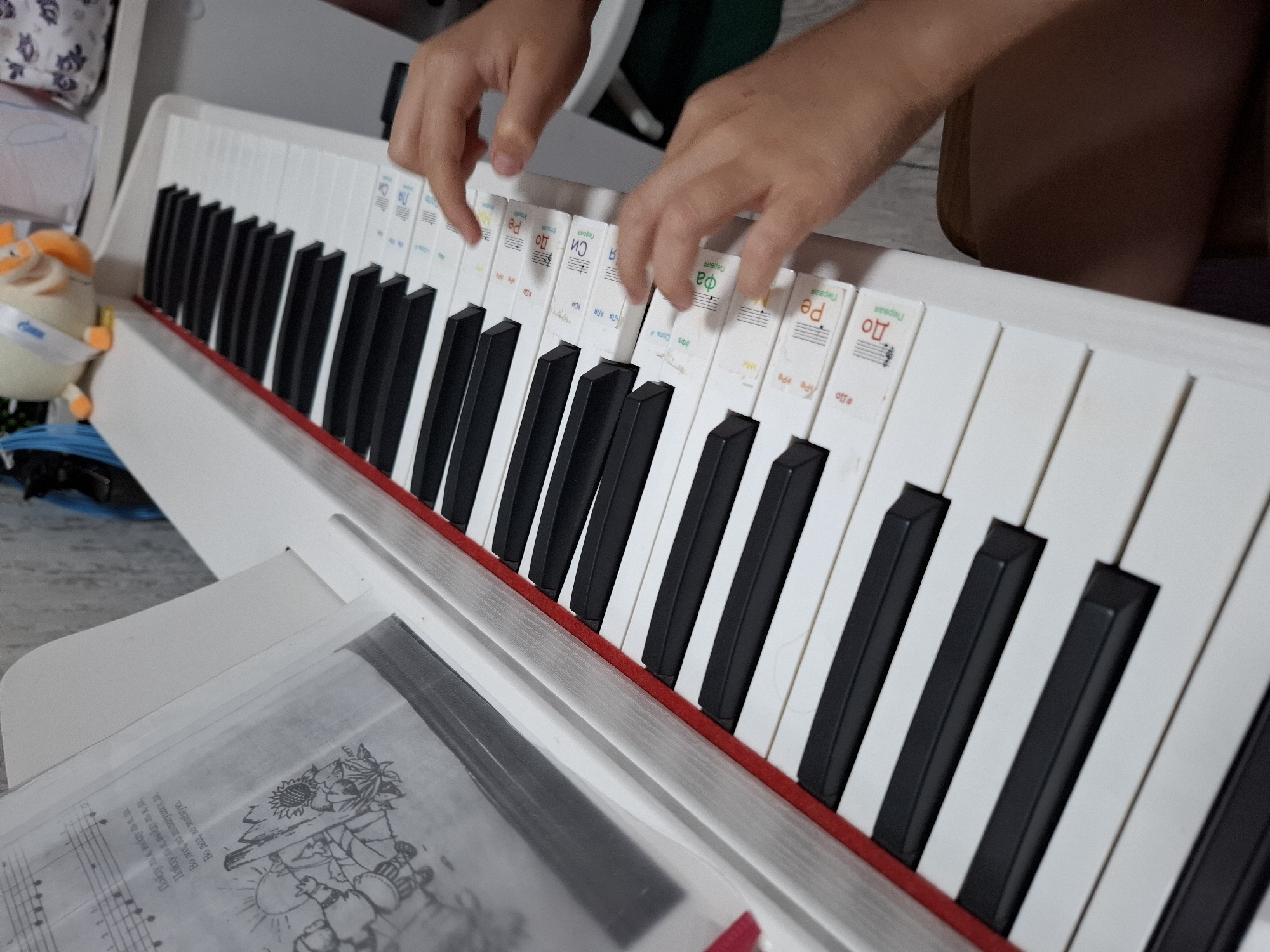 The image shows a close-up view of a white electronic piano with black keys. A sheet of music with Cyrillic text and an illustration is propped up on the music stand. Two hands are positioned over the keys, with fingers poised to play. The keys have small, colorful stickers with Cyrillic musical notation and notes. The background is blurred, but appears to be an indoor setting with a wall and possibly some clutter. There is no visible indication of the city of Korenovsk, Russia in the image.