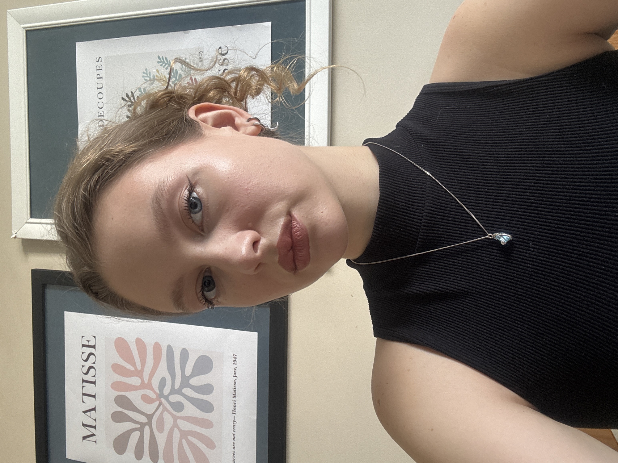 The image shows a young woman with blonde hair pulled back, wearing a black sleeveless top and a silver necklace with a blue pendant. She has blue eyes and light makeup. In the background, there are two framed art prints on a light-colored wall. One print, to the left, features the word "MATISSE" and an abstract floral design in pink, grey, and white. The other print, to the right, has the word "DECOUPES" visible and a partial floral illustration with green accents. The lighting suggests an indoor setting, likely a room. The image is a close-up, upper-body portrait of the woman, taken from a slightly low angle. There is no information available to indicate the location in the city of Izhevsk, Russia.