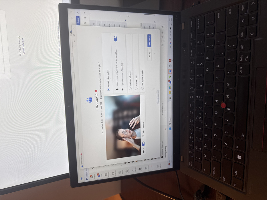 The image depicts a black Lenovo ThinkPad laptop, featuring a Polish keyboard layout with a red TrackPoint, situated on a dark brown surface. The laptop screen displays the Microsoft Teams application, open to a pre-meeting setup for a meeting titled "LOVE SQUAD ❤️". The meeting is scheduled for "czwartek, 8 sty" (Thursday, January 8th), from "10:00 - 11:00 CET" (Central European Time), which corresponds to Warsaw, Poland. The interface indicates "Organizator + Inne osoby, które dołączyły: 2" (Organizer + Other people who joined: 2).
A video preview window within Teams shows a young woman with glasses, dark hair tied back, wearing a white shirt and a dark blazer. She is holding a smartphone in her left hand, looking at its screen, with her right hand near her temple. The background behind her is blurred. An audio settings pop-up is active, with "Dźwięk z komputera" (Sound from computer) selected, and both "Microphone Array (Intel® Smart Sound Tec...)" and "Speakers (Realtek(R) Audio)" enabled. Options for "Dźwięk z telefonu" (Sound from phone), "Dźwięk z sali" (Sound from room), and "Nie używaj dźwięku" (Do not use sound) are visible but not selected. Buttons "Anuluj" (Cancel) and "Dołącz teraz" (Join now) are present.
The Windows 11 taskbar is visible at the bottom of the laptop screen, showing the current time as 10:01 and the date as 08.01.2026 (January 8, 2026). Above and behind the laptop, a secondary monitor is partially visible, displaying a webpage, likely a Microsoft service, with the text "Don't have the app? Download it now." The overall environment appears to be an indoor setting with subdued lighting.