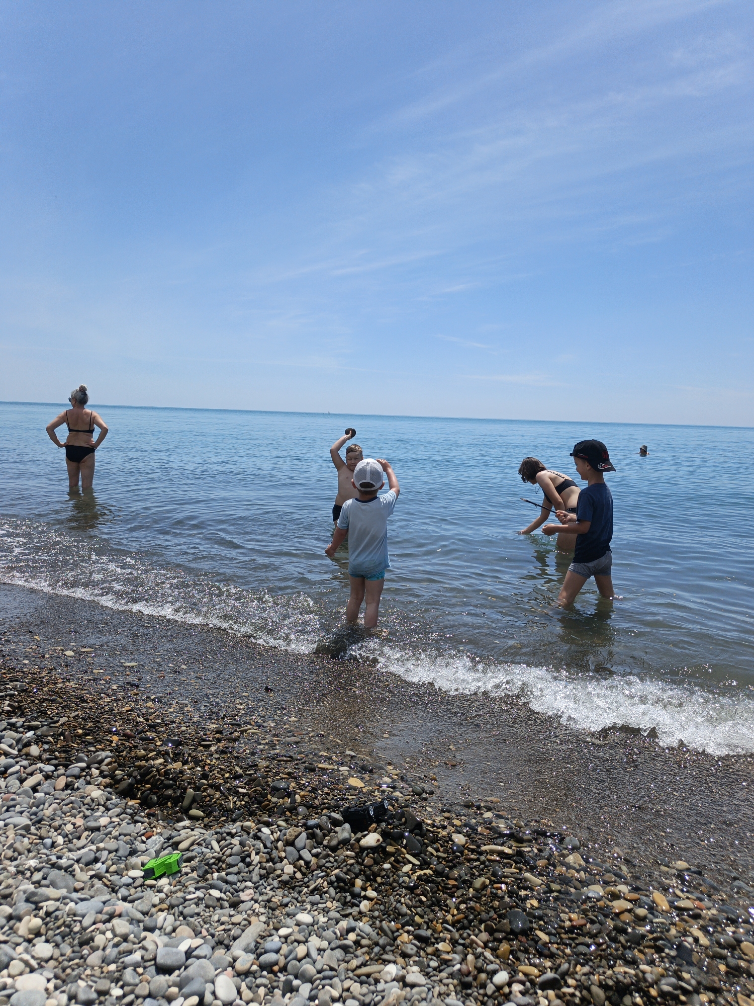 This image depicts a sunny day at a pebbled beach in Krinitsa, Russia. The sky is clear light blue with minimal, faint wisps of white clouds. The calm, blue-green sea extends to the horizon, where two dark, vertical posts are visible further out in the water on the right side. Small waves are breaking softly against the shore, creating a wet line on the beach composed of numerous grey, brown, and white pebbles.
Five individuals are present in the shallow water near the shore. On the far left, an adult woman with grey hair, wearing a black bikini, stands with her back to the viewer, hands on her hips, looking out at the sea. Her lower thighs are submerged. To her right, a young child wearing a light-colored t-shirt and light blue shorts with a white baseball cap worn backward stands facing away from the viewer, submerged up to the knees.
In the center, another child, shirtless and wearing dark swim trunks, faces the viewer while holding a dark, possibly rocky object aloft with both hands. This child is submerged approximately to the waist. To their right, a third child, wearing a dark swimsuit, is bent over, appearing to search or interact with the water, holding a thin, elongated object like a stick. This child is also submerged to the waist. On the far right, a fourth child, wearing a dark blue t-shirt and grey patterned swim trunks with a dark baseball cap worn forward, stands facing the viewer, submerged up to the knees.
On the pebbled beach in the foreground, a small, bright green plastic toy is visible.