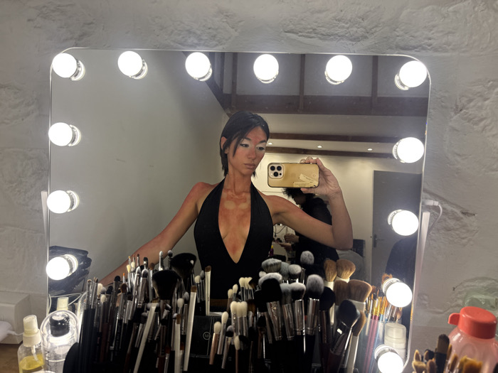 The image displays a woman taking a mirror selfie in a well-lit vanity setting. The central subject, reflected in a large rectangular mirror framed by 12 illuminated spherical bulbs, is a woman with short, dark hair, wearing a black halter-neck top. Her chest, shoulders, and arms are covered in an orange-red application with several distinct, uncolored circular patches. Her face features prominent white makeup around her eyes and on portions of her forehead and cheeks. She holds a gold-colored smartphone in her raised right hand, capturing her reflection.
In the immediate foreground, a dense collection of makeup brushes, varying in size and type, is arranged upright in multiple containers on a surface below the mirror. Adjacent to these, several bottles, including clear ones containing liquids and a red-capped plastic container, are visible.
The mirror's reflection also reveals the room's interior, characterized by whitewashed, textured stone or brick walls and exposed wooden ceiling beams. Recessed lights are visible on the ceiling. A second person with dark, textured hair is partially visible in the background reflection, positioned behind the main subject's arm and phone. A grey door with a handle is also reflected in the background. The scene is located in a studio or preparation room in Paris, France.
