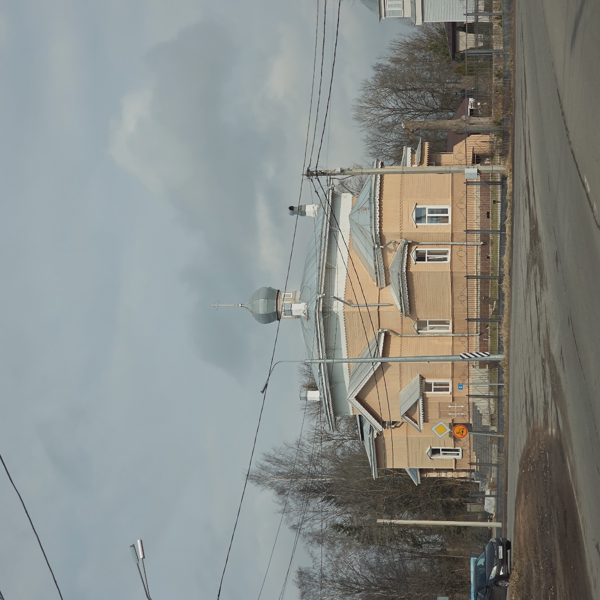 The image depicts a street scene in Volosovo, Russia, featuring a wooden church with a silver dome and a cross. The church is tan-colored and has multiple windows. A metal fence surrounds the property. Power lines are visible across the sky, intersecting with the church's dome. A road runs alongside the church, with a car parked at the edge. A road sign with a yellow diamond and a restriction of 5 tons is visible near the church. The sky is overcast. Bare trees are present in the background.