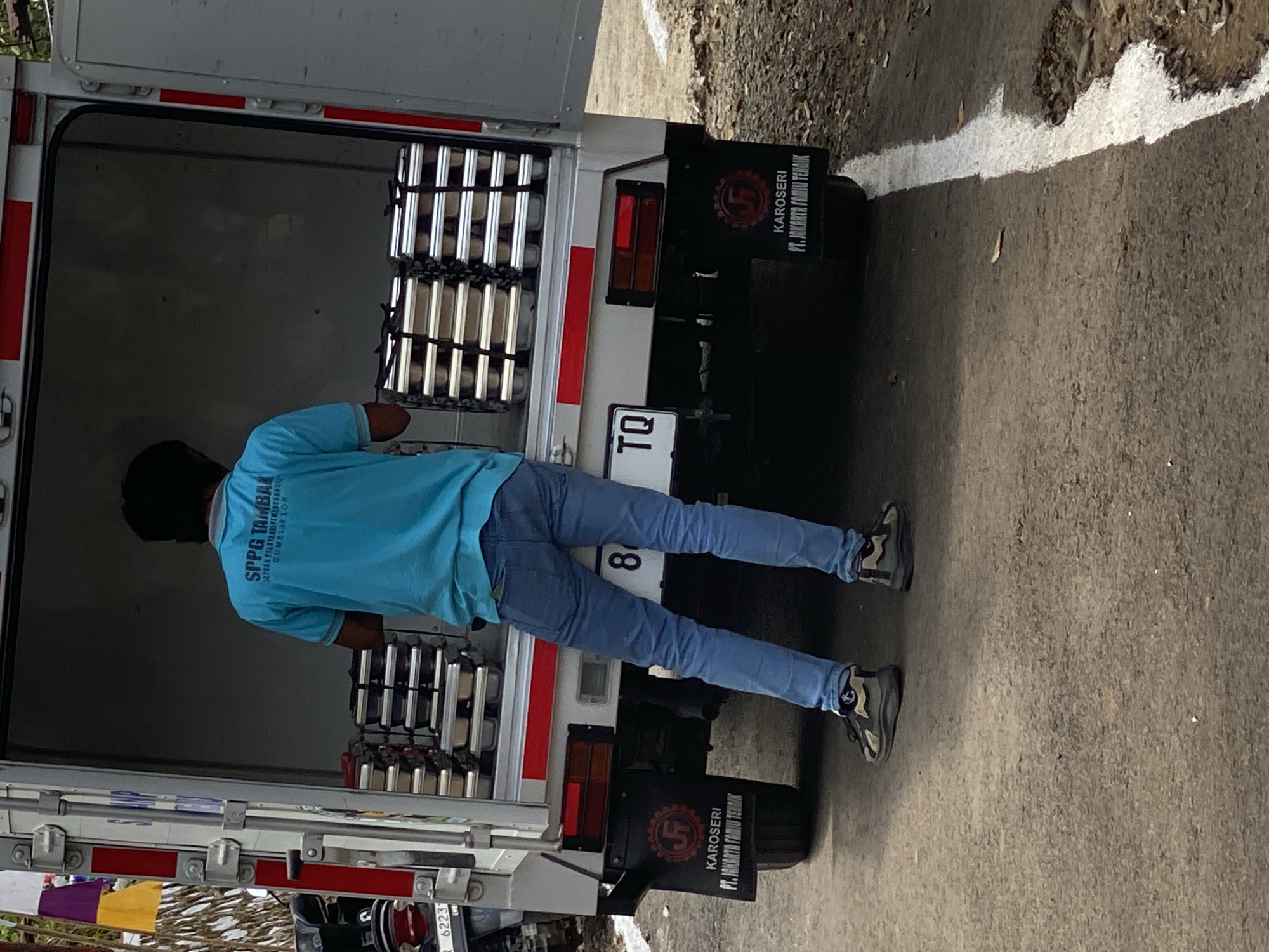 A man in a blue t-shirt and jeans is standing at the back of a white truck, with the rear doors open. He is facing away from the camera, looking into the truck bed where several stacks of metal containers are visible. The truck has a license plate that reads "8 TQ" and a logo on the rear fender that says "KAROSERI PT. JAKARTA FAMILI TEMBAK". The scene appears to be on a paved road in Gumelar Kidul, Indonesia.