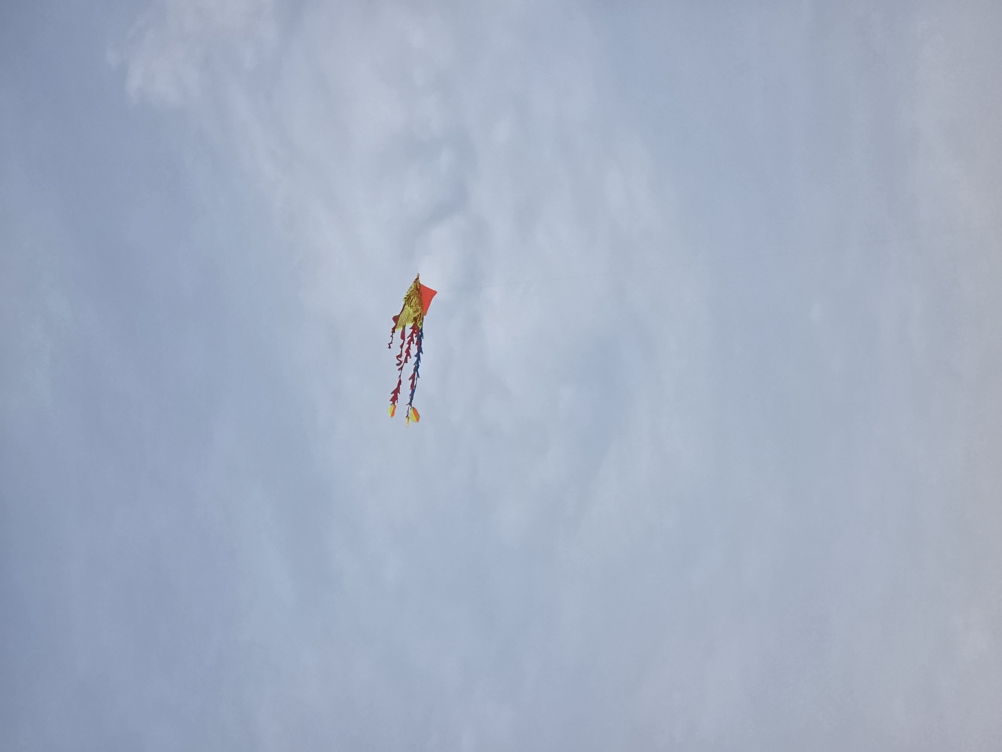 A colorful dragon kite is flying in a cloudy sky. The kite is predominantly yellow and orange with red and blue streamers trailing behind it. The kite is positioned in the upper right quadrant of the image. The sky is overcast with soft, diffused clouds, suggesting a slightly hazy or overcast day. There are no people or other objects visible in the frame. The image appears to be taken from a ground-level perspective, looking upwards. The location of Đà Nẵng, Vietnam, is not evident from the visual information within the image itself.
