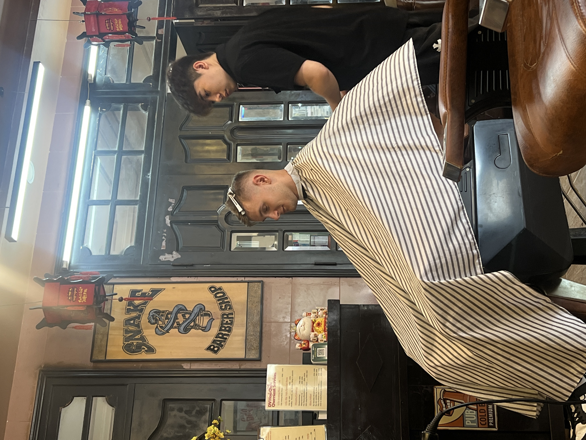 The image captures a scene inside the "Snake Barber Shop" in Bắc Nha Trang, Vietnam. A barber in a black shirt is attending to a customer who is draped in a black and white striped cape. The barber is leaning over the seated customer, who has a clip in their hair. Behind them, there is a wall decoration with the words "Snake Barber Shop" on it. There is also an advertising sign with text beginning with "PUB". In the background is a window.