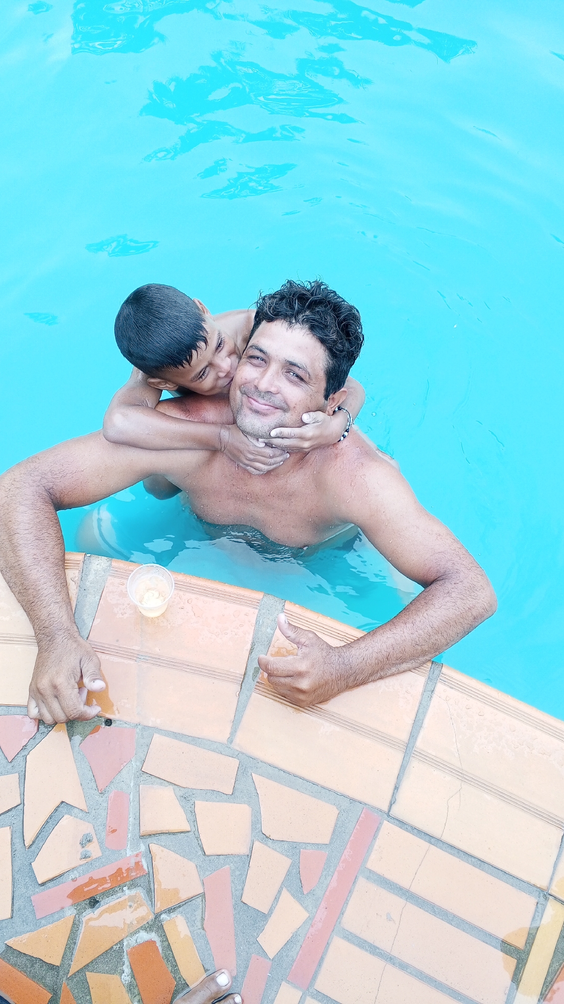 The photograph shows a man and a boy in a swimming pool. The boy is clinging to the man's back and neck. The man is leaning against the pool's edge, which is tiled in various shades of orange and red. There is a small cup containing a yellow substance on the pool's edge. The location is Unknown, Unknown.