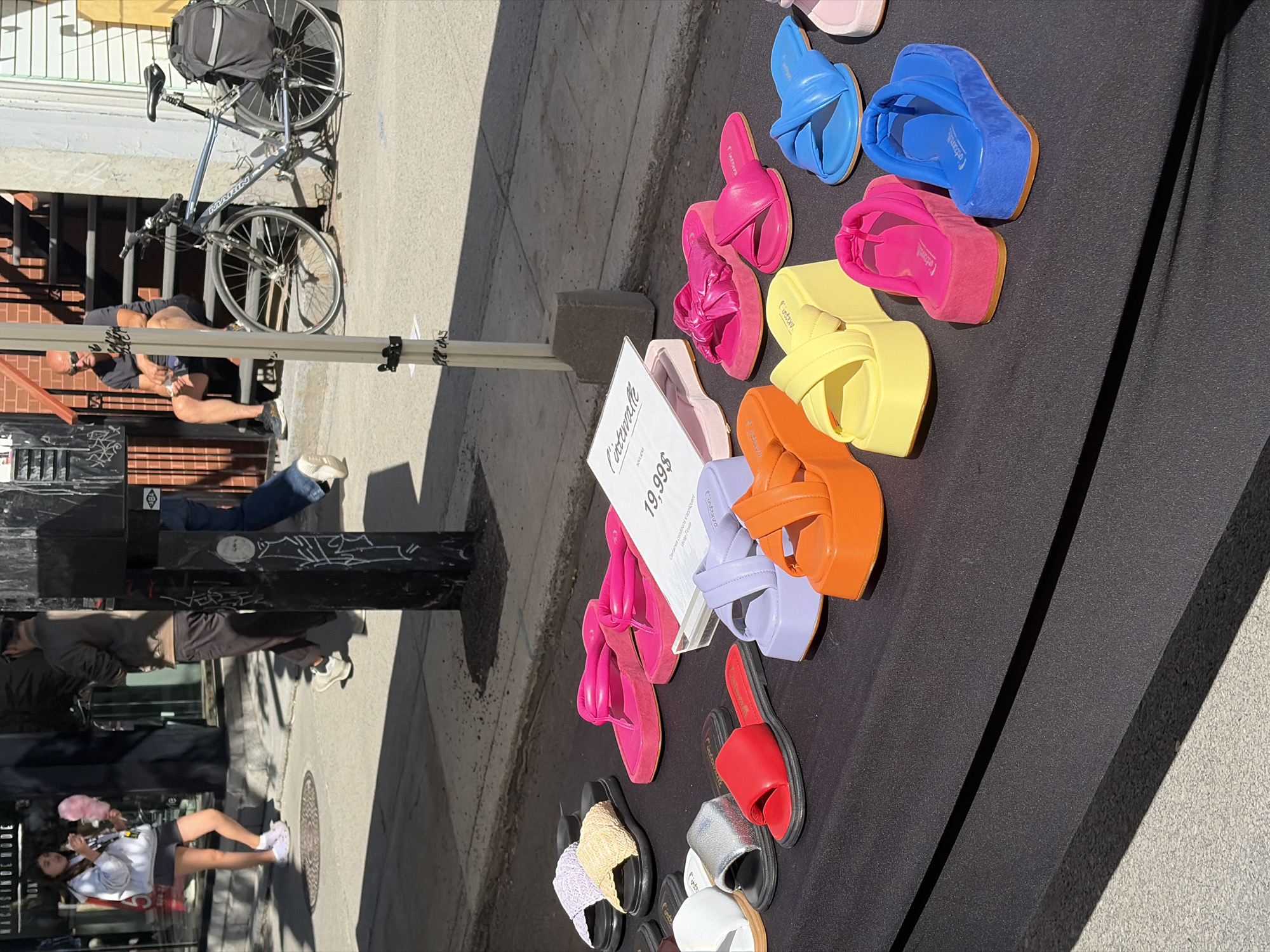 The image shows a variety of colorful platform sandals displayed on a black surface outdoors, likely for sale. A sign indicates a price of $19.99. In the background, there is a street scene, including a bicycle, pedestrians, and storefronts, suggesting the location is in a commercial area of Montreal, Canada. There are reflections on the storefront in the bottom-left of the picture.