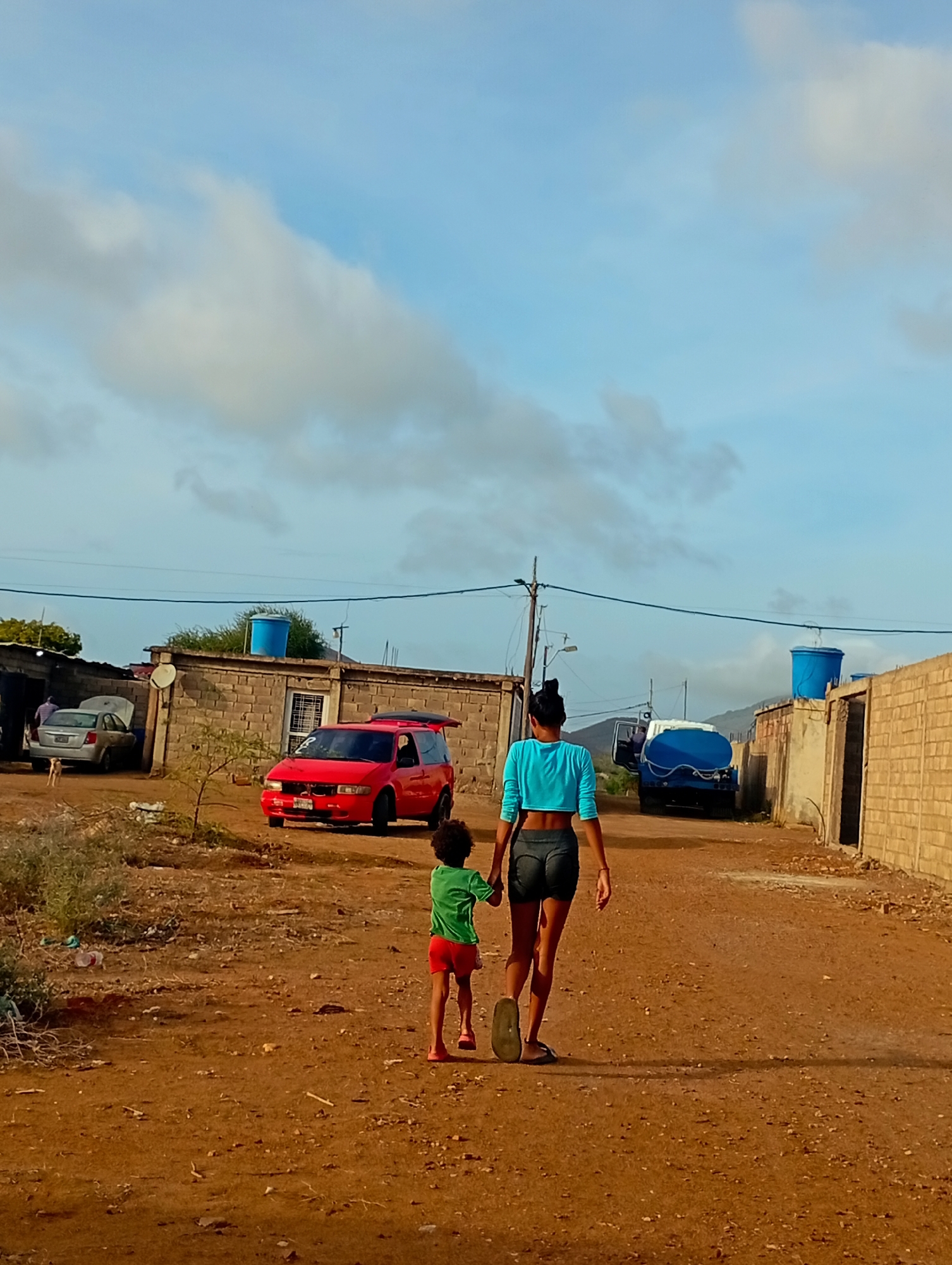 In Altagracia, Venezuela, under a cloudy sky, a woman and a child walk away from the camera on a dirt road. The woman wears a teal crop top and dark shorts, while the child is dressed in a green t-shirt and red shorts. They are in a semi-arid environment with low-lying buildings constructed from concrete blocks. A red minivan is parked on the left side of the road, and further down, a blue water tanker truck is visible. The ground is covered in dirt and sparse vegetation.