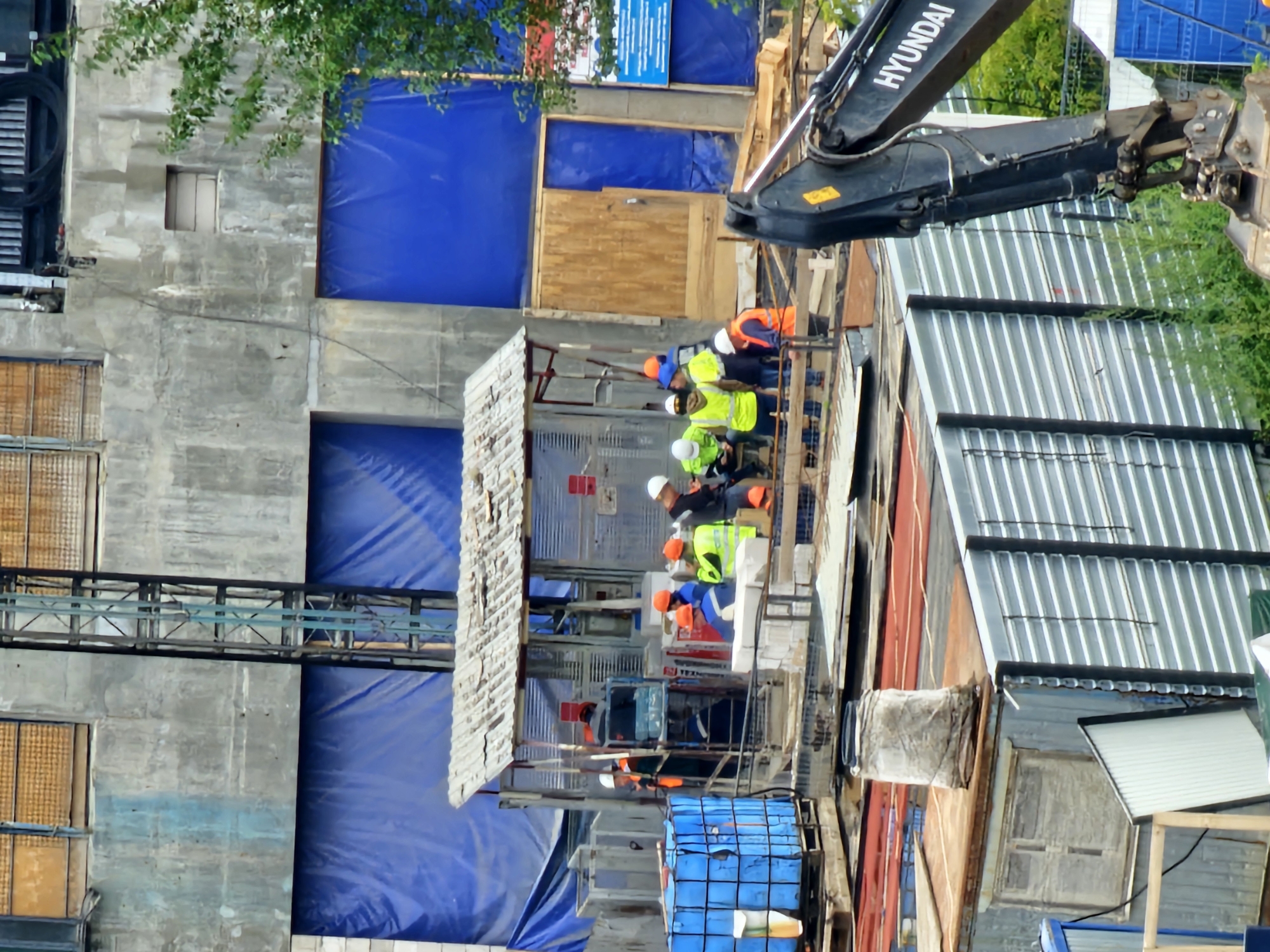 The image depicts a construction site with several workers. The scene features a partially constructed building with blue tarpaulin covering large sections. A metallic structure, possibly a hoist or elevator shaft, is visible on the left side of the building. In the foreground, there are stacks of building materials, including pallets with blue containers and other construction supplies. A small, makeshift shelter with a corrugated roof is also present.
Several individuals, all wearing hard hats and some in high-visibility vests, are engaged in various activities. Some are standing and conversing, while others appear to be working or observing. A large excavator, branded with "HYUNDAI," is positioned in the lower right corner of the image, its arm extended. The environment suggests an active construction zone.
While the image itself does not contain explicit markers of location, the presence of Cyrillic text on a blue sign in the upper right quadrant suggests the location is in Russia. Given the provided context, this construction site is identified as being in Moscow, Russia.