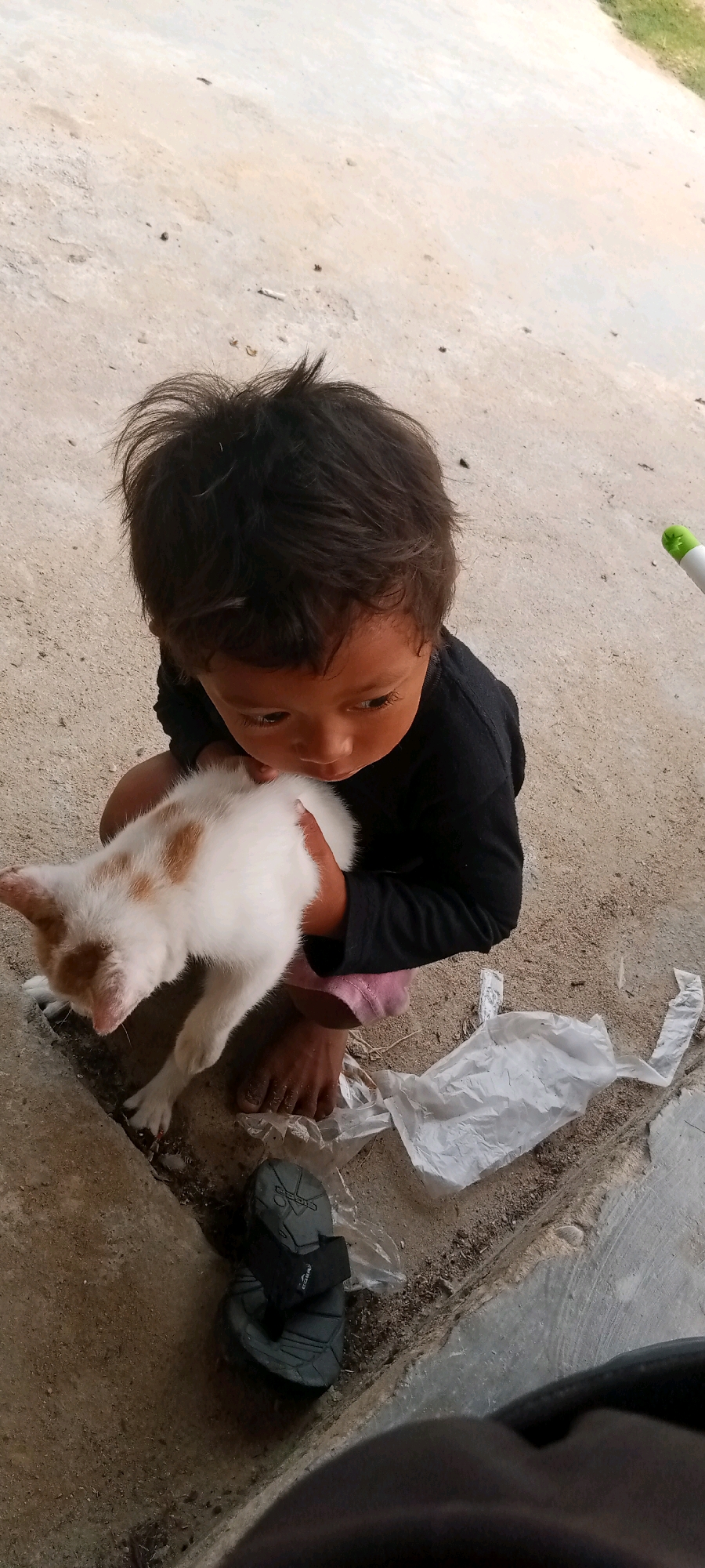 In Kerumutan, Indonesia, a young child with dark, tousled hair kneels on a dirt and concrete surface, holding a white and ginger kitten. The child wears a black long-sleeved shirt and pink shorts. The kitten is held close to the child's chest. In the foreground, a gray sandal lies on the ground next to a crumpled piece of white plastic. A green and white object, possibly a pen or marker, is visible in the upper right portion of the frame. The environment appears to be an outdoor area with a rough, uneven ground surface.