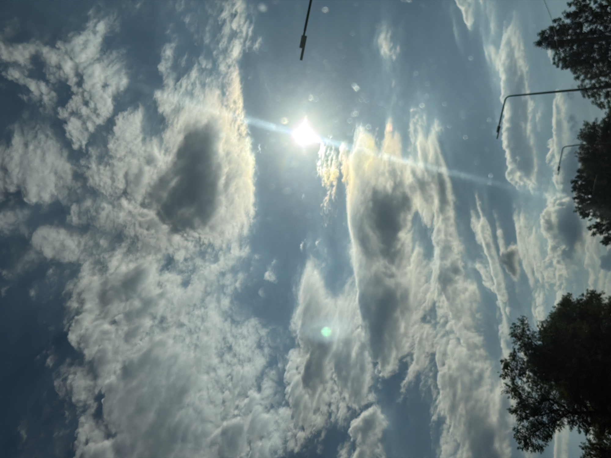 The image depicts a daytime outdoor scene dominated by a cloudy sky with the sun partially obscured. There are several streetlights visible, along with the tops of trees in the foreground. The sky is a mix of blue and grey, with the sun creating bright lens flares and a visible light beam. The overall impression is of a partially overcast but bright day. The location is identified as Teofipilka, Ukraine.