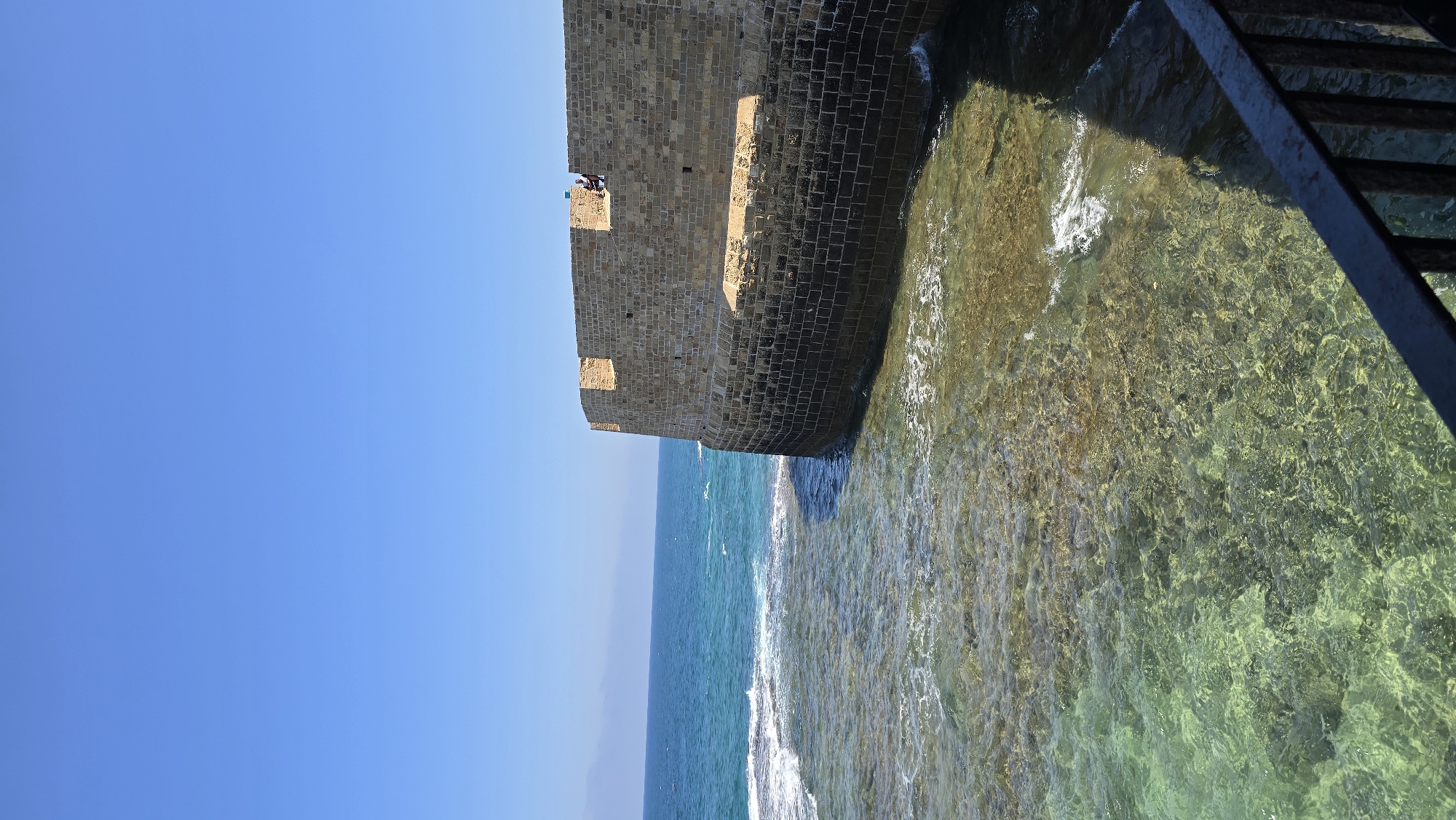 A wide-angle, eye-level, outdoor, daytime shot captures a section of the ancient sea walls of Acre, Israel, bordering the Mediterranean Sea. The massive, curved fortification, constructed of rough-hewn, light-brown stone blocks, dominates the right side of the frame, casting a dark shadow onto the clear, shallow water at its base. The upper portions of the wall are lighter in color, with indentations and crenellations, suggesting a historical defensive structure. Two figures, small in the frame and seated near the top of the wall, are visible, one appearing to be in white. The sea itself is a vibrant turquoise, with small waves gently lapping against the base of the wall and breaking over submerged rocks or sandbars, creating patches of white foam. The water is remarkably clear, revealing the textured seabed beneath. The sky is a solid, cloudless, bright blue, meeting the horizon in the distance. A dark, metal railing runs across the bottom of the frame, partially obscuring the foreground. The overall impression is one of historical grandeur against a backdrop of a tranquil, sun-drenched seascape.