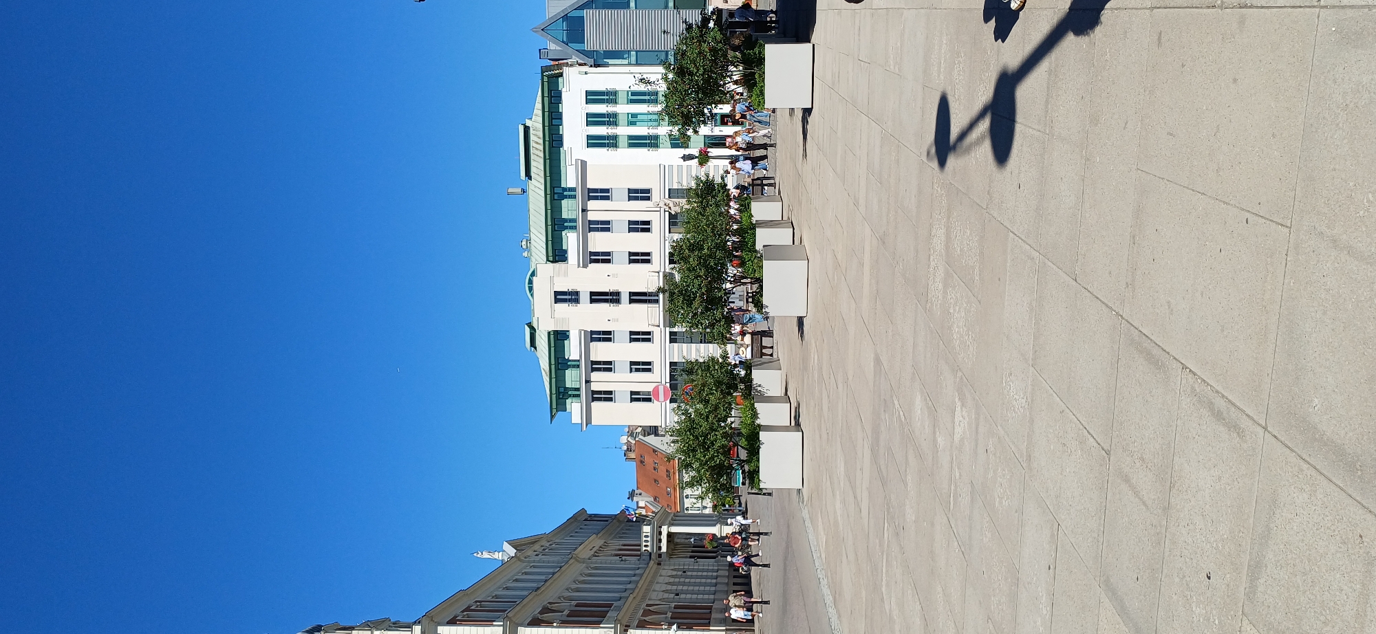 This media file is an outdoor photograph taken on a sunny day, likely in the late morning or early afternoon given the shadows. The scene depicts a wide, paved plaza with several modern buildings lining one side. The buildings are a mix of white and light-colored facades with numerous windows. Some of the buildings have green roofs and architectural details such as balconies or overhangs. Trees are planted in large square planters along the edge of the plaza, providing some greenery.
Several people are visible in the image, mostly walking or sitting at outdoor tables near the buildings, suggesting a public space like a cafe or restaurant. Their attire suggests a casual, warm-weather setting. The shadows cast by a streetlamp and the people indicate the bright sunlight. The sky is a clear, vibrant blue.
The perspective of the image is somewhat tilted, as if the photographer was holding the camera at an angle or the scene itself is on an incline. The plaza appears clean and well-maintained. There is no visible text in the image. The overall impression is one of a pleasant day in a European-style urban environment.