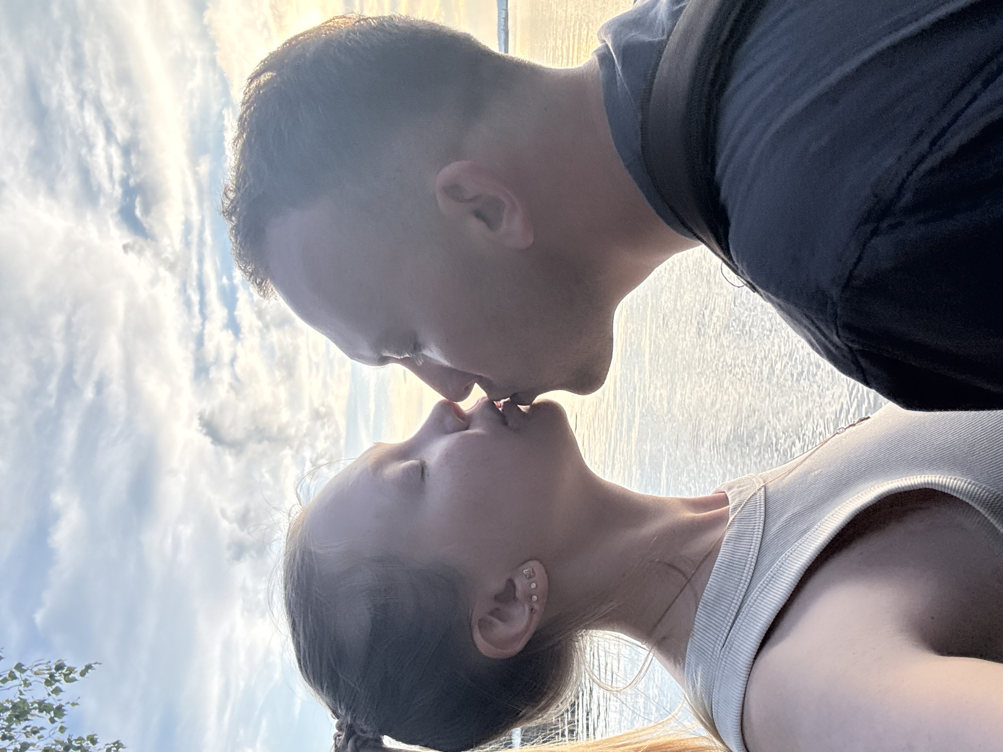 An outdoor medium shot shows a young couple kissing with their faces in profile. The woman is on the left, and the man is on the right. The woman has her eyes closed and is wearing a light-colored tank top and a necklace. She has blonde hair in a braid and several earrings in her left ear. The man is wearing a dark t-shirt and has short hair. They are positioned in front of a body of water that reflects the sky, which is filled with clouds and hints of sunlight. The scene appears to be taking place outdoors, possibly near a waterfront, given the presence of water and a sky with scattered clouds. The overall lighting suggests it is either dawn or dusk.