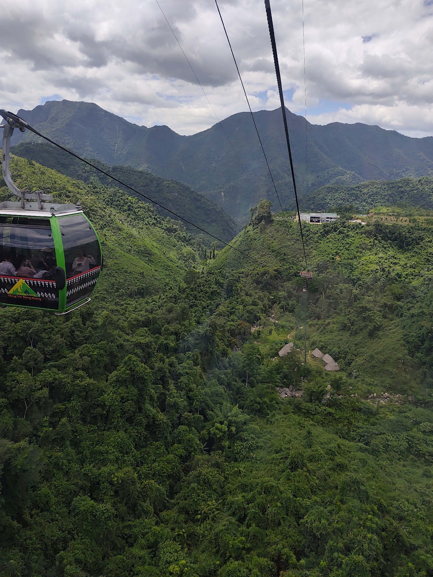 A green and black cable car, with several passengers visible inside, travels along a thick black cable suspended above a lush, densely forested mountainside. The cable car is on the left side of the frame and is angled upwards. In the distance, more mountains are visible, shrouded in a hazy, cloudy sky. A modern building complex with white walls and dark roofs is situated on a distant hill. The overall scene depicts a scenic mountain landscape with a cable car system, suggesting a tourist attraction.