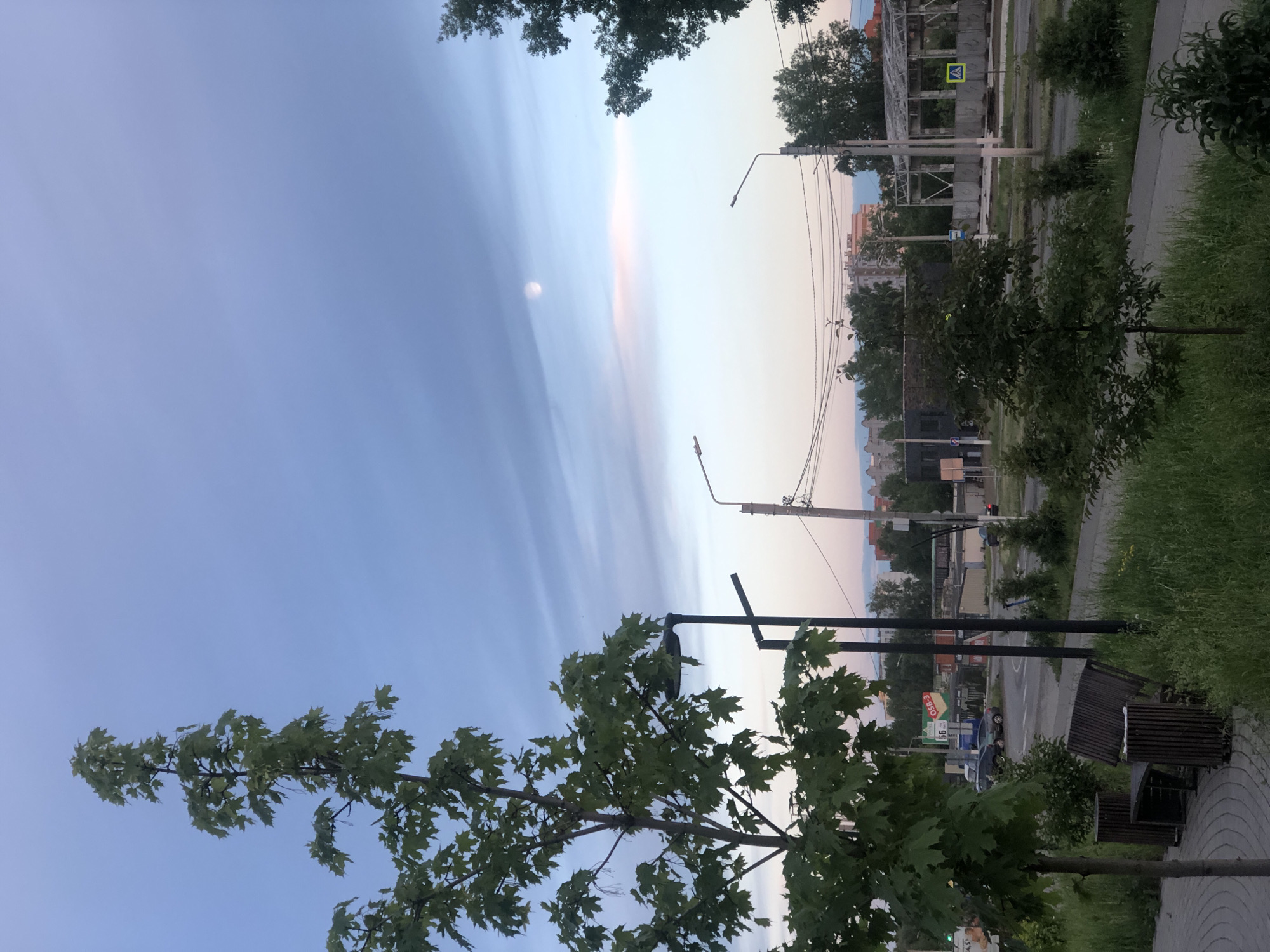 The image captures a view in Chaikovsky, Russia, at dusk or dawn. In the foreground, there are trees and benches. A sidewalk runs along the right side of the frame, adjacent to a street. Several lampposts are visible. In the background is the skyline of the city, with buildings and utility poles. The sky is streaked with colors, showing blue and pink hues. There is a full moon visible in the sky. A pedestrian crossing sign is visible.