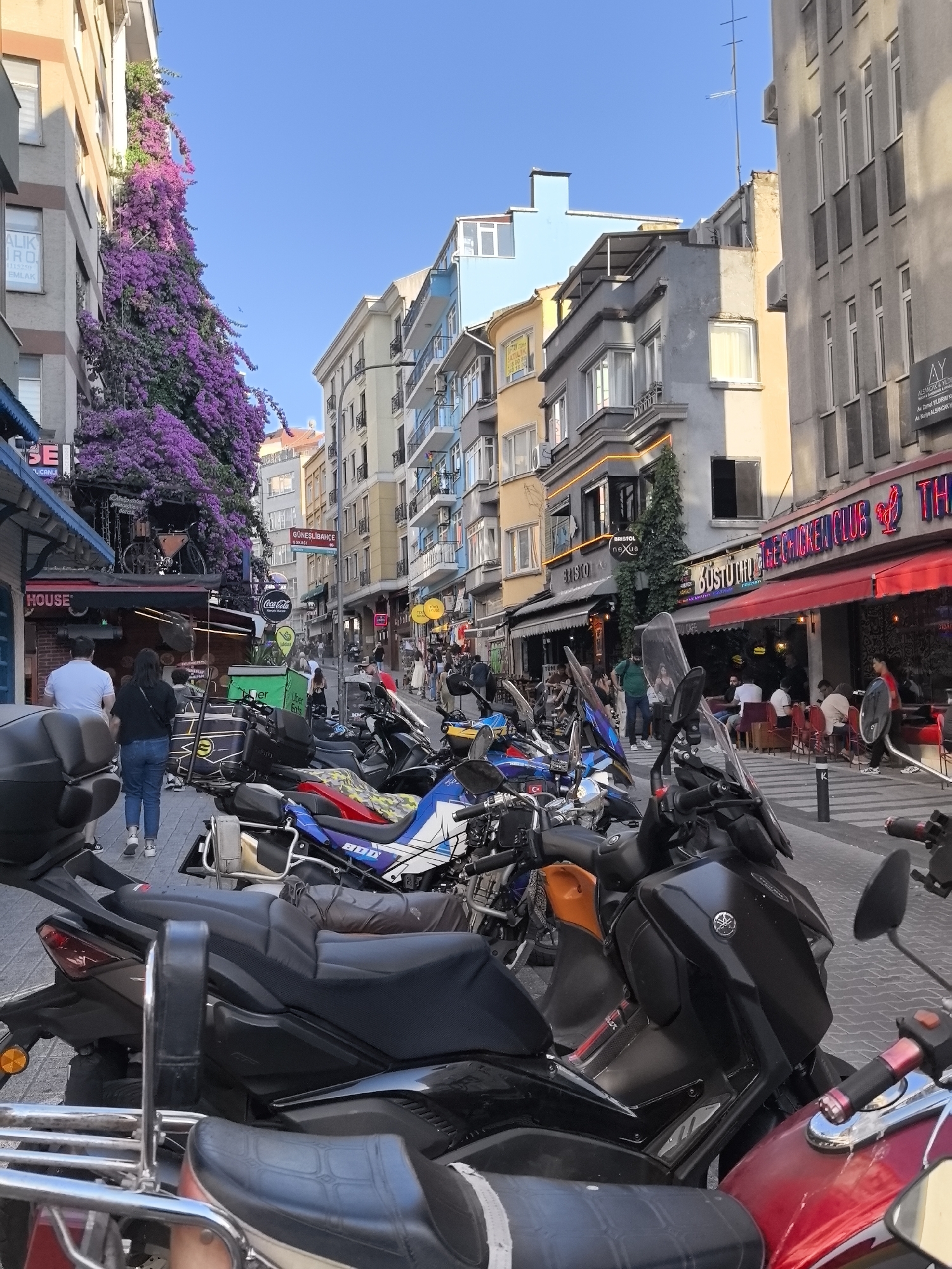 This is a vertical, eye-level, outdoor shot of a street in Kadıköy, Turkey. The scene is bustling with activity, likely in the late afternoon or early evening, given the warm, golden light casting long shadows. The street is lined with multi-story buildings housing various businesses, including restaurants and shops. Brightly colored signs adorn the storefronts, with visible text like "Coca-Cola," "Uber Eats," and "The Chicken Club" suggesting a lively commercial district.
The foreground is dominated by a row of parked motorcycles, their sleek forms and various colors adding to the urban feel. Beyond the motorcycles, pedestrians are seen walking along the sidewalk, some entering or exiting establishments. Bougainvillea flowers, a vibrant cascade of purple, spill down the side of one building, providing a striking contrast to the urban architecture and hinting at a Mediterranean climate. The sky is a clear, bright blue, indicating pleasant weather. The overall impression is one of a vibrant, lively urban environment, full of life and commerce.