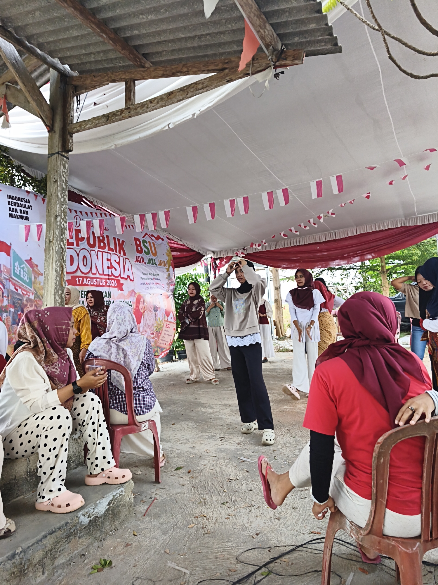 The image shows an outdoor gathering under a canopy supported by wooden beams. The canopy is decorated with red and white bunting. In the background, a large banner reads "PUBLIK BSU INDONESIA 17 AGUSTUS 2026". Several people are present, mostly women wearing hijabs. Some are seated on chairs or steps, while others are standing. One woman in a light-colored sweatshirt and black pants is in the center, appearing to be performing or interacting with others. Another woman in a red shirt and maroon hijab is seated facing away from the camera, with her arm resting on the back of a chair. The ground is paved with concrete and scattered leaves. The overall scene suggests a community event or celebration. Based on the banner text, the event is likely related to Indonesia's Independence Day celebrations in 2026. Without specific landmarks, pinpointing the exact location within Bandar Lampung is not possible, but it appears to be an open-air public space or community center.