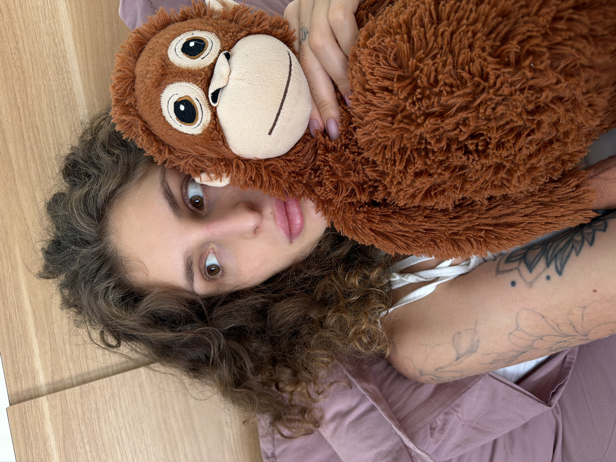 A woman with curly brown hair lies on a bed covered in a light purple sheet. She holds a brown plush monkey toy close to her face. The woman has fair skin, brown eyes, and pink lips. Her fingernails are painted light pink. A tattoo is visible on her left arm. A light-colored wooden surface is partially visible at the edge of the frame. The location, North Kuta, Indonesia, cannot be determined from the image content alone.