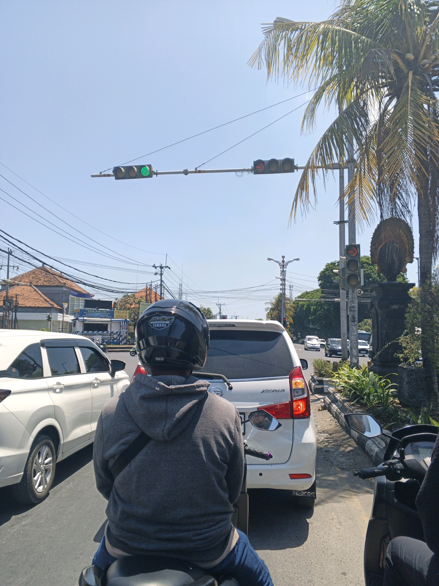 This image shows a street scene in Denpasar, Indonesia. The perspective is from the back of a motorcycle, looking forward at traffic and the surrounding environment.
In the foreground, a person wearing a gray hoodie and a black helmet is seated on a motorcycle, facing forward. The motorcycle's handlebars and part of the seat are visible. To the left of this motorcycle, a white car is stopped in traffic, showing its side profile. To the right, another motorcycle is partially visible, with its handlebars and part of the rider's leg in view.
The road surface is asphalt. The traffic appears to be stopped, likely at an intersection, as evidenced by the traffic lights. There are multiple traffic lights visible. One traffic light, suspended over the intersection, shows a green light. Another traffic light, mounted on a pole to the right of the intersection and near a palm tree, shows a red light and a red arrow pointing to the right.
The environment includes a clear blue sky. A tall palm tree with prominent fronds is on the right side of the image. Behind the palm tree, there is a stone sculpture or statue, partially obscured by foliage. On the left side of the street, buildings with orange tile roofs are visible, as well as power lines crisscrossing overhead. There are also some signs on the buildings, including one that appears to be for "POLISI" (police). The overall impression is a typical busy urban street scene in a tropical climate.