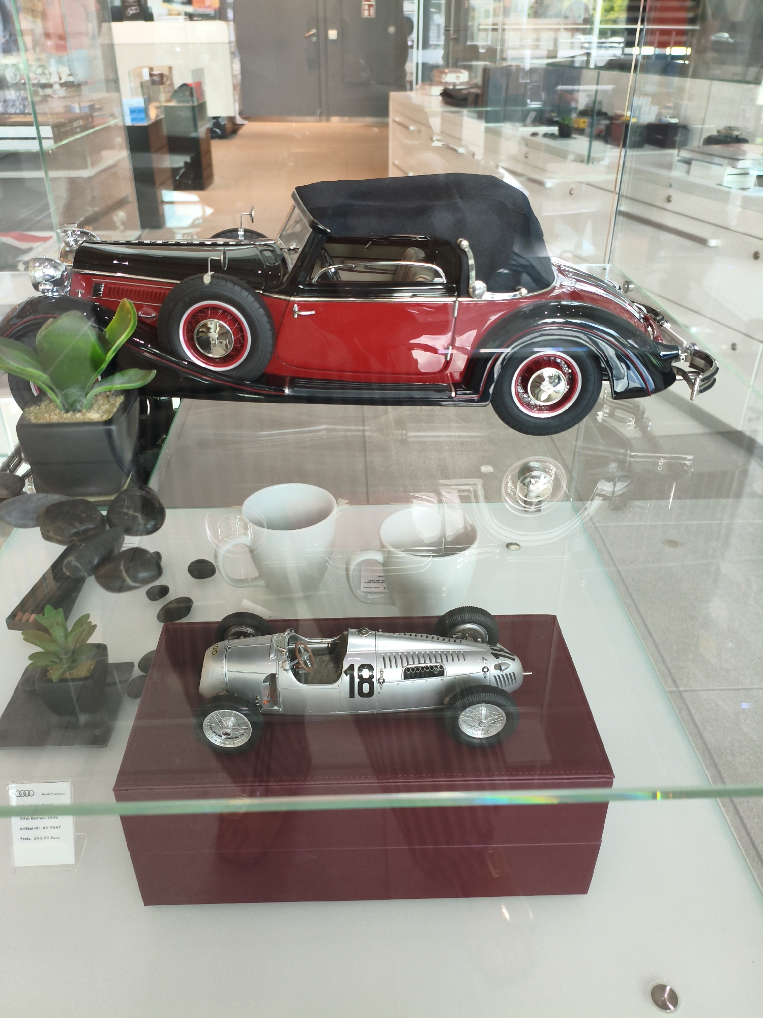 The image shows a display case in what appears to be a museum or showroom, likely the Audi Forum in Ingolstadt, Germany. Inside the display case are two highly detailed model cars. The upper model is a classic red and black convertible with its top down, featuring a spare tire mounted on the side. Below it, on a maroon-colored box, sits a silver vintage race car with the number "18" prominently displayed on its side. The display case also contains decorative elements such as potted plants and smooth stones. A label is visible on the lower left, providing details about the silver race car, including "Eifel Rennen 1936," "Artikel-Nr. AS-5897," and "Preis: 392,00 Euro." The overall environment suggests a clean and well-lit retail or exhibition space. There are no people visible in the image.