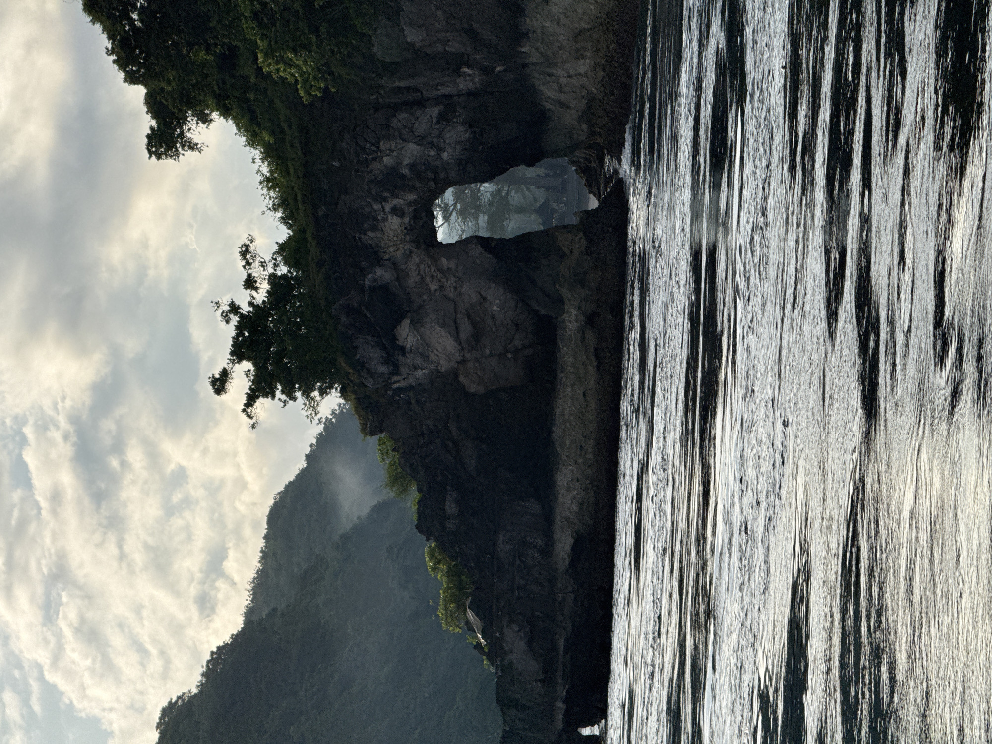 This image shows a partially submerged cliff face with a natural archway, likely in Nusa Penida, Indonesia. The lower portion of the image displays water reflecting the sky. The cliff face is dark and rocky, with some vegetation on top. The archway reveals more land beyond it. The sky is filled with clouds.