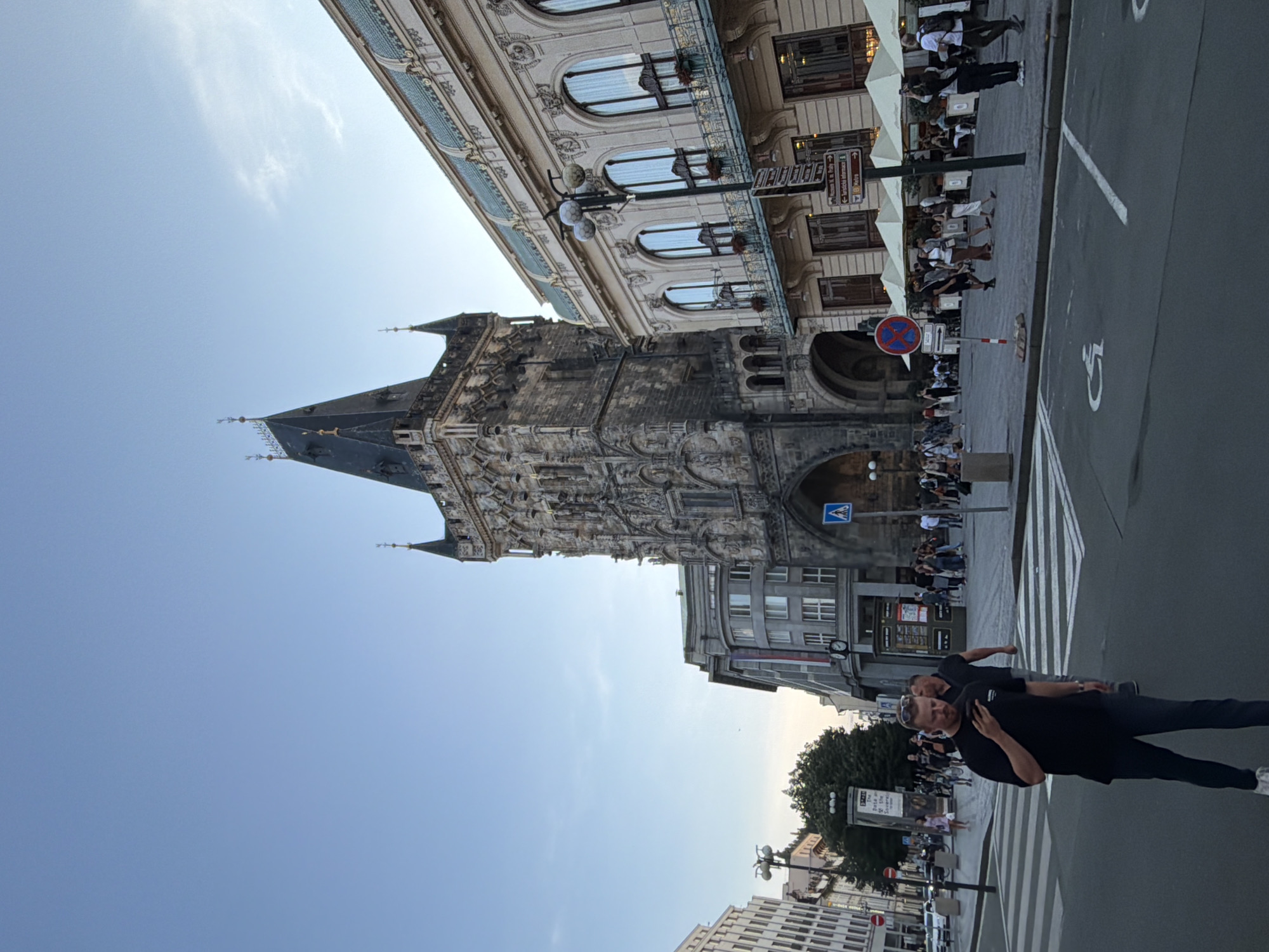 The image shows a wide, grey street in Prague, Czechia, with a tall, gothic tower dominating the background on the right. The tower features intricate stone carvings and a dark, pointed roof. To the right of the tower is a grand, ornate building with large arched windows and a cream-colored facade.
In the foreground, a man in a black t-shirt and dark pants stands on a pedestrian crossing, looking at his phone. He is the most prominent human figure. Behind him, other pedestrians are visible, some walking and others gathered near outdoor seating areas under umbrellas. A few cars are parked on the side of the street, and traffic lights and street signs are present. The sky is a clear, pale blue. The overall scene suggests a bustling urban environment in the late afternoon or early evening.