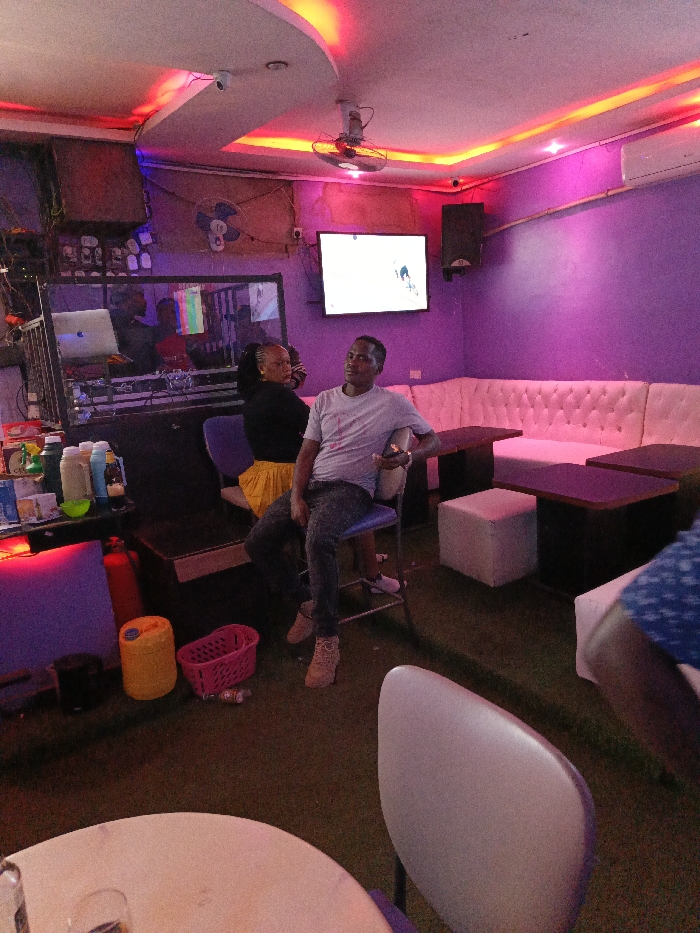 This image displays an interior view of an entertainment establishment, likely a bar or lounge, located in Nairobi, Kenya. The room features purple-painted walls with red and orange ambient LED cove lighting integrated into a dropped ceiling. A wall-mounted fan and an air conditioning unit are visible. The floor is covered in green material, resembling artificial turf or carpet.
On the right side, an L-shaped white tufted bench seating arrangement with multiple dark, rectangular tables is present. White square poufs are situated as auxiliary seating or footrests. In the foreground, a partial view of a round table and a white chair is observed.
Towards the center-left, a male subject is seated on a light blue chair, facing slightly to his left while holding a mobile phone in his right hand. He wears a light grey t-shirt, dark jeans, and light brown boots. Behind him, a female subject is seated on a similar light blue chair, facing away from the camera, adorned in a black top and a bright yellow skirt. Her hair is styled in braids. A third individual, wearing a blue and white patterned shirt, is partially visible on the far right.
On the left, a DJ booth enclosed by glass or reflective panels contains a person in a red shirt operating a MacBook Pro laptop and other DJ equipment. Below the booth, a counter holds various bottles and a green bowl. On the floor adjacent to the counter, a red fire extinguisher, a yellow plastic jerrycan, and a pink plastic basket are positioned.
A flat-screen television mounted on the right wall displays what appears to be a sports event. A security camera (CCTV) is affixed to the ceiling, and a speaker is mounted on the wall beneath the air conditioning unit. The specific location within Nairobi is not discernible from the visual information provided.