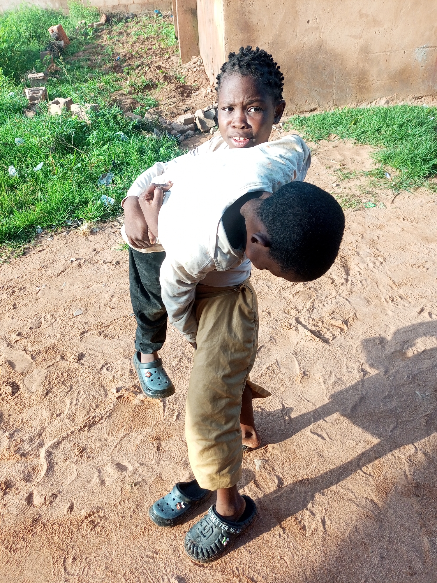 In Tempure, Nigeria, two children are shown outdoors. One child, facing the camera, is wearing a light-colored long-sleeved shirt and black pants, with dark gray clogs. The other child, facing away from the camera, is wearing a similar light-colored long-sleeved shirt and light brown pants, also with dark gray clogs. The child facing away is holding the child facing the camera. They are standing on a sandy surface. In the background, there is dry grass and a brick wall. The scene is illuminated by sunlight, casting shadows.