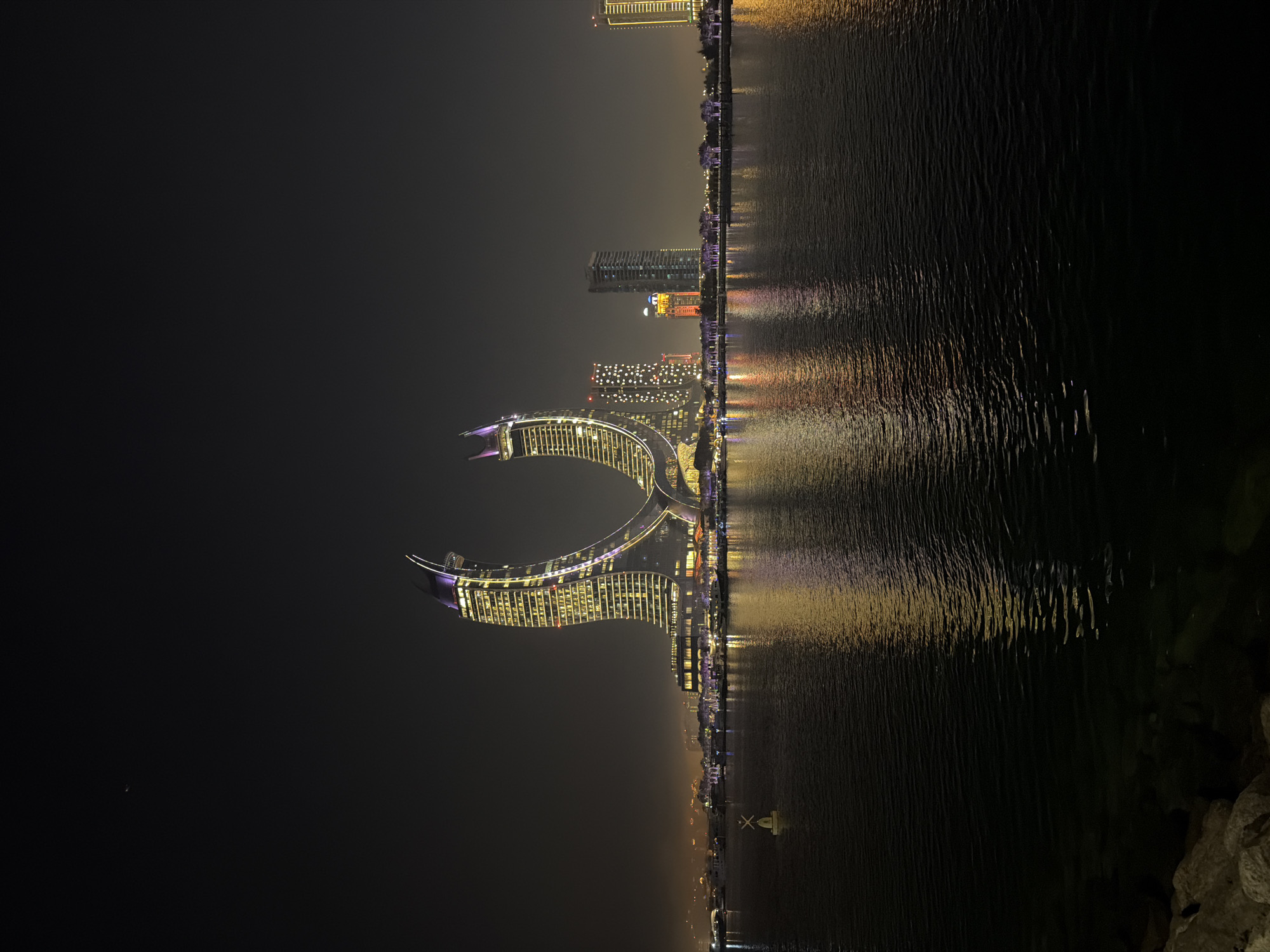 This is a nighttime, landscape-oriented photograph of a waterfront cityscape. The primary subject is a crescent-shaped building illuminated with yellow and purple lights, which reflects on the dark water in front of it. The building appears to be a modern architectural marvel, possibly a hotel or residential complex, with many windows visible.
Beyond this prominent structure, a skyline of other illuminated buildings stretches across the horizon. These buildings vary in height and design, with some featuring unique patterns of lights. The water's surface is rippled, creating a distorted but colorful reflection of the city lights, suggesting a calm but not entirely still body of water. The overall atmosphere is one of urban vibrancy and nighttime beauty.
The time of day is clearly night, and the weather appears to be clear, with no visible precipitation. The lighting from the buildings is the main source of illumination, casting a warm glow on the scene. The location is a waterfront, likely a city known for its modern architecture and developed coastline. The mood evoked is one of awe and wonder at the illuminated cityscape. No people are visible in the frame, and there is no visible text in the image. The photograph's orientation is rotated 90 degrees clockwise.