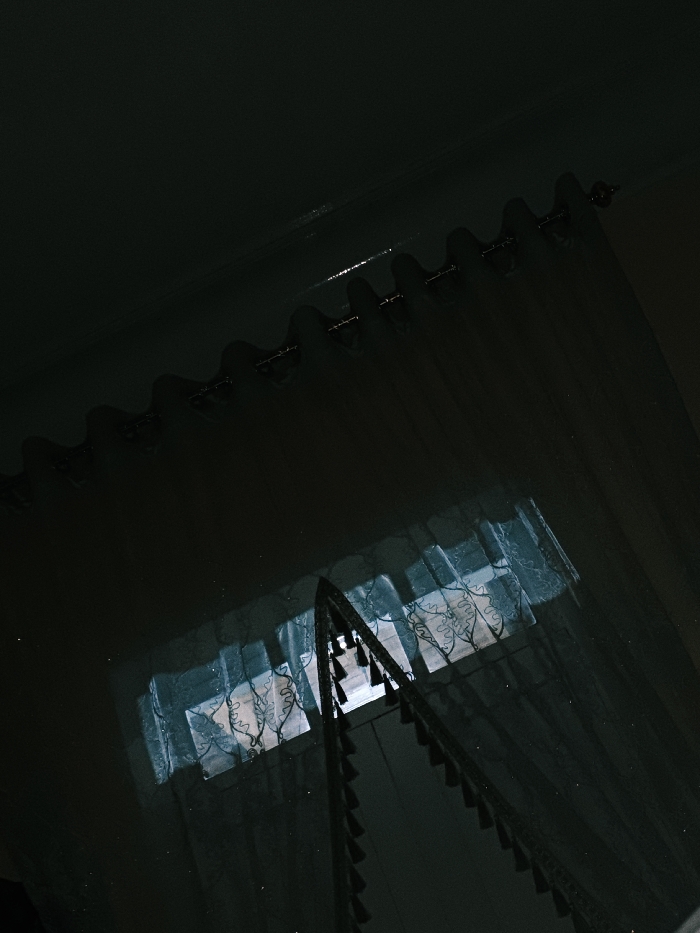 A low-angle shot showcases a window covered by sheer curtains. Sunlight filters through the window and curtains. The curtain rod and rings are visible at the top, with evenly spaced scallops in the curtain fabric. A decorative valance with hanging tassels adorns the window. The room is dimly lit, emphasizing the contrast between the bright window and the dark surroundings.