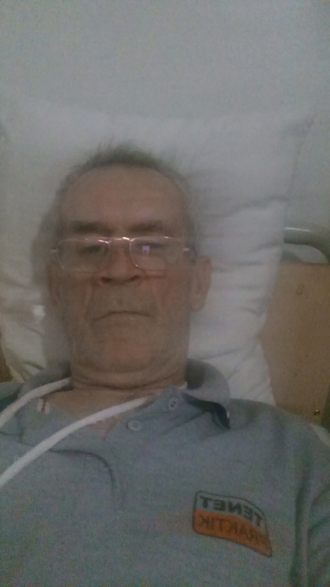 A middle-aged man with graying hair and glasses is lying in a hospital bed, taking a selfie. He is wearing a light gray polo shirt with a logo on the left side of his chest. A white tube is visible around his neck. The background is blurry and appears to be a hospital room, with a white pillow and a portion of a bed railing visible. The lighting is dim. There is no identifiable information in the image that would indicate the location in the city of Tiachiv, Ukraine.