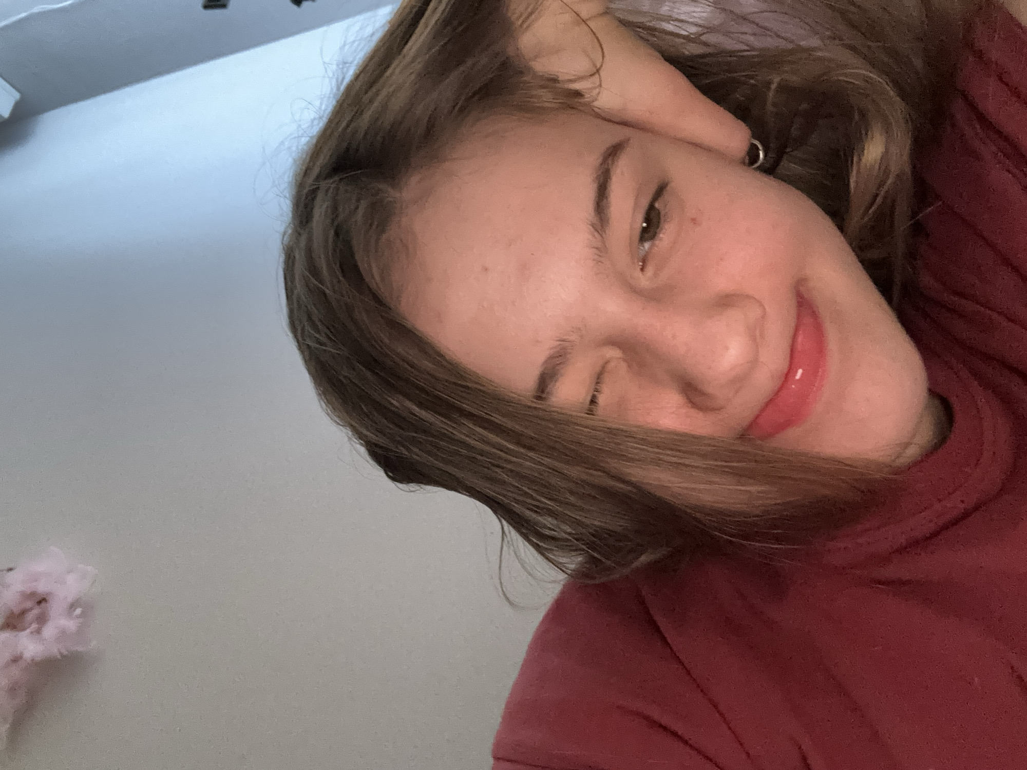 A young person is pictured indoors, leaning their head on their hand and winking. They have brown hair and are wearing a maroon shirt. The background is a plain wall. There is a pink fluffy object in the upper left corner of the image. The image was taken in Vsevolozhsk, Russia.
